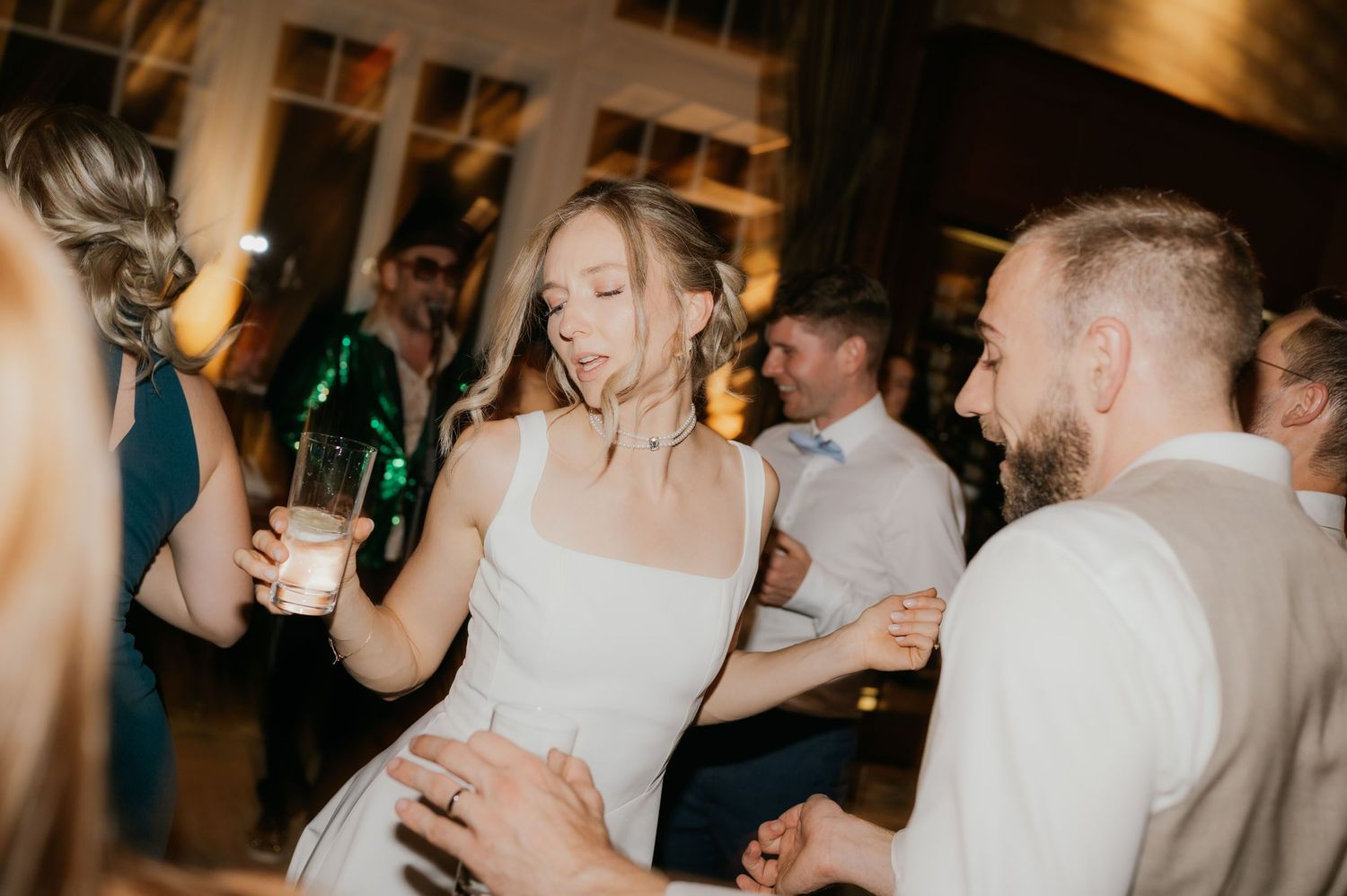 Bride in white dress dancing with guests at wedding reception, holding drink and smiling joyfully.