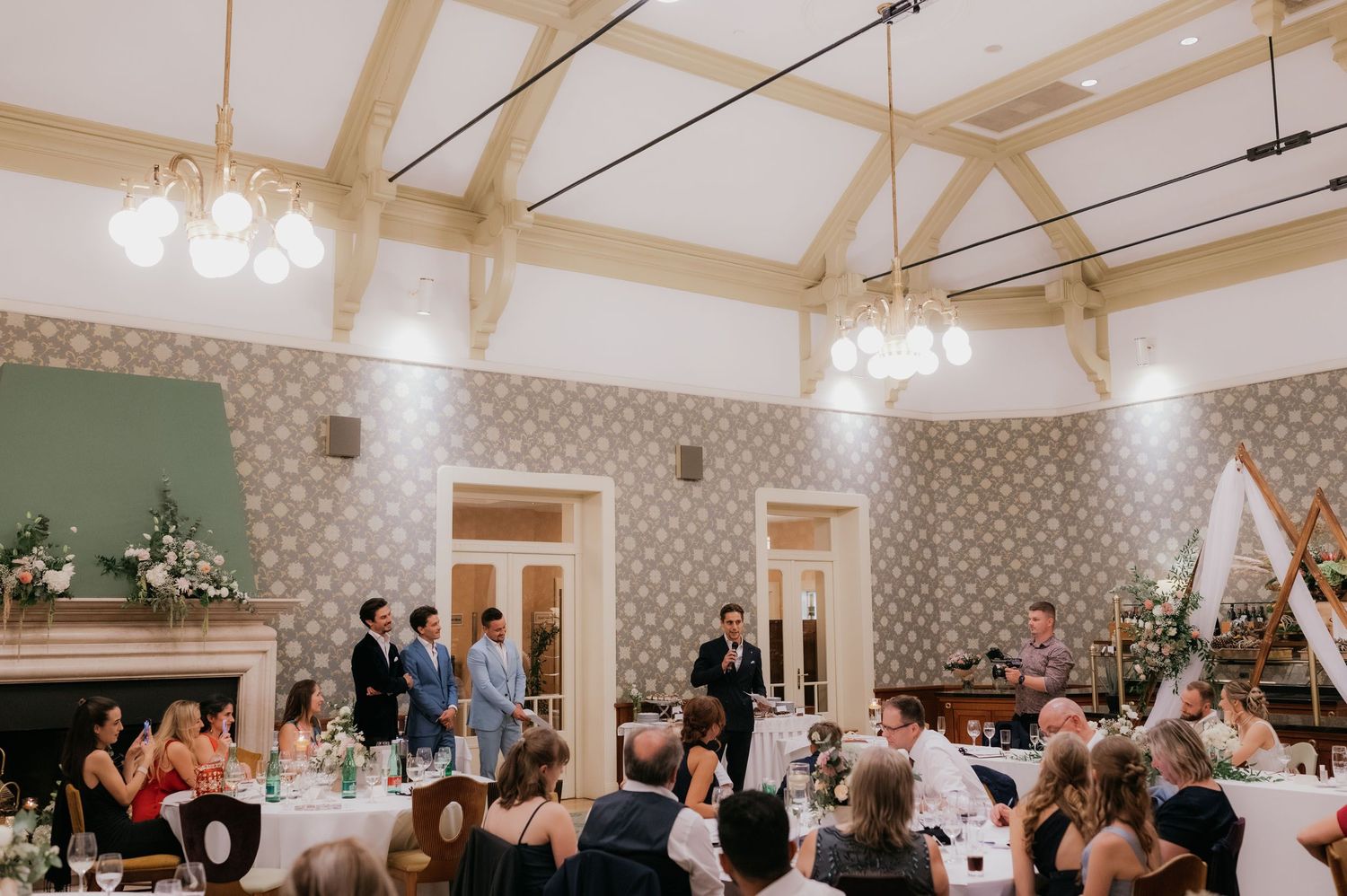 Elegant wedding reception in a decorated hall with chandeliers, floral centerpieces, and guests seated at round tables.