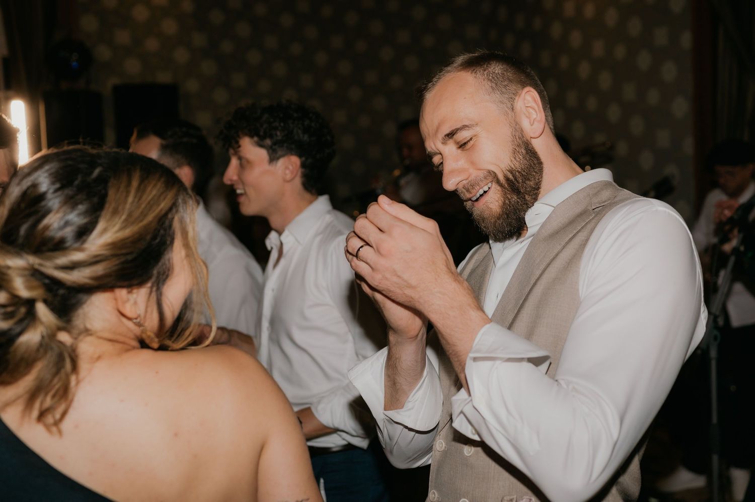 Groom smiling while dancing with guest at wedding reception, wearing vest and white shirt.
