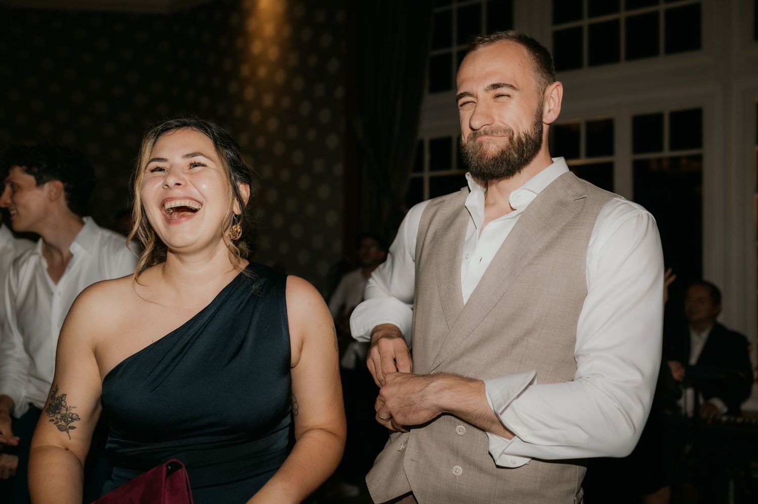 Laughing couple at wedding reception, woman in dark dress with tattoo, man in beige vest and white shirt.