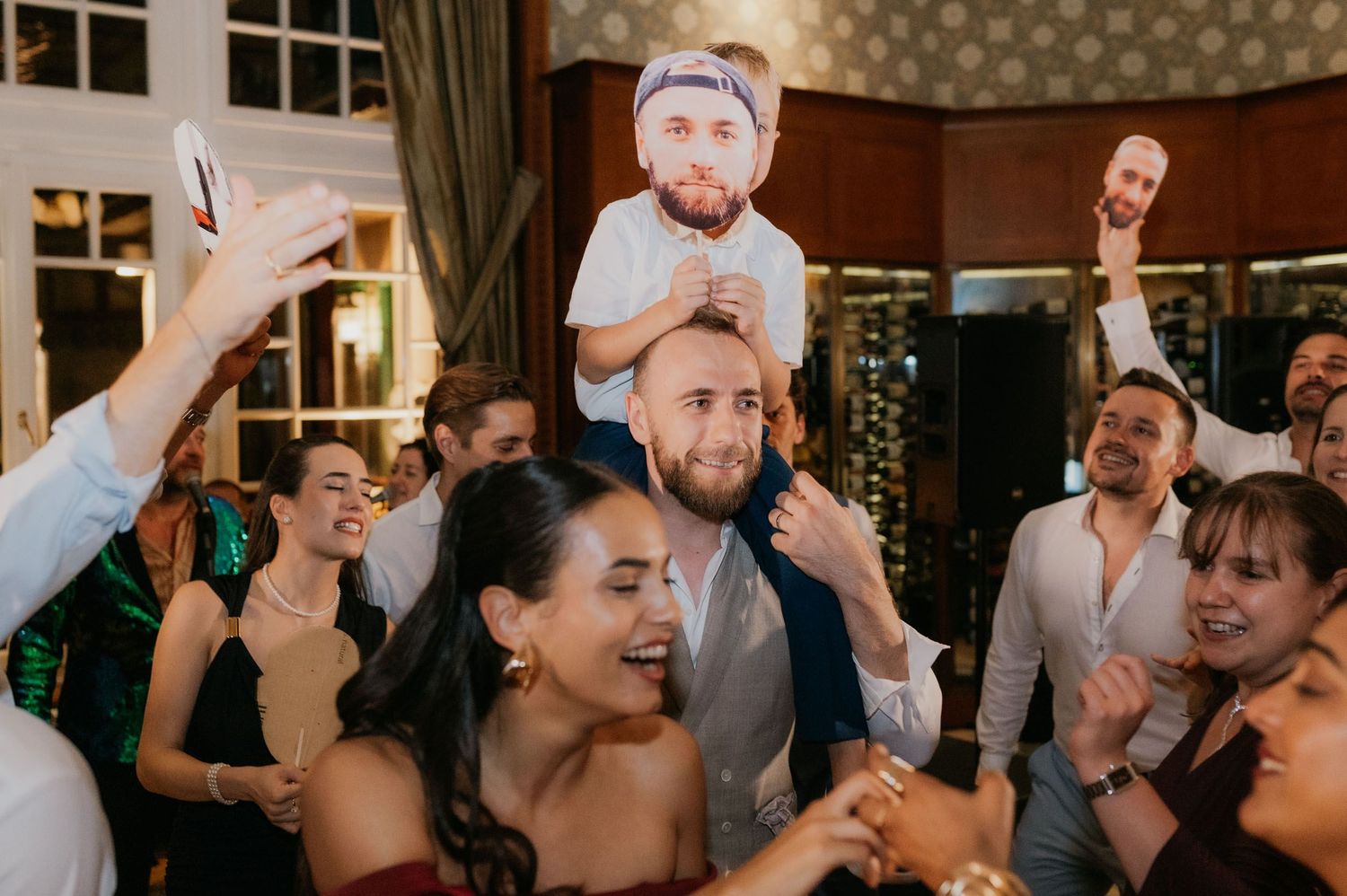 Wedding guests celebrate on dance floor, laughing and holding oversized face cutouts at a lively indoor reception party.
