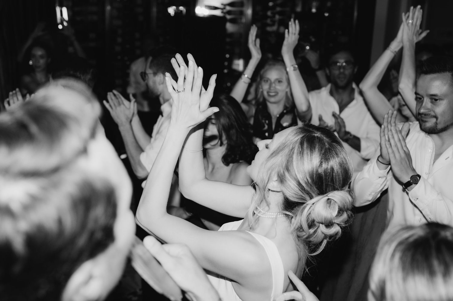Black and white photo of wedding guests dancing with hands raised at a lively wedding reception celebration.