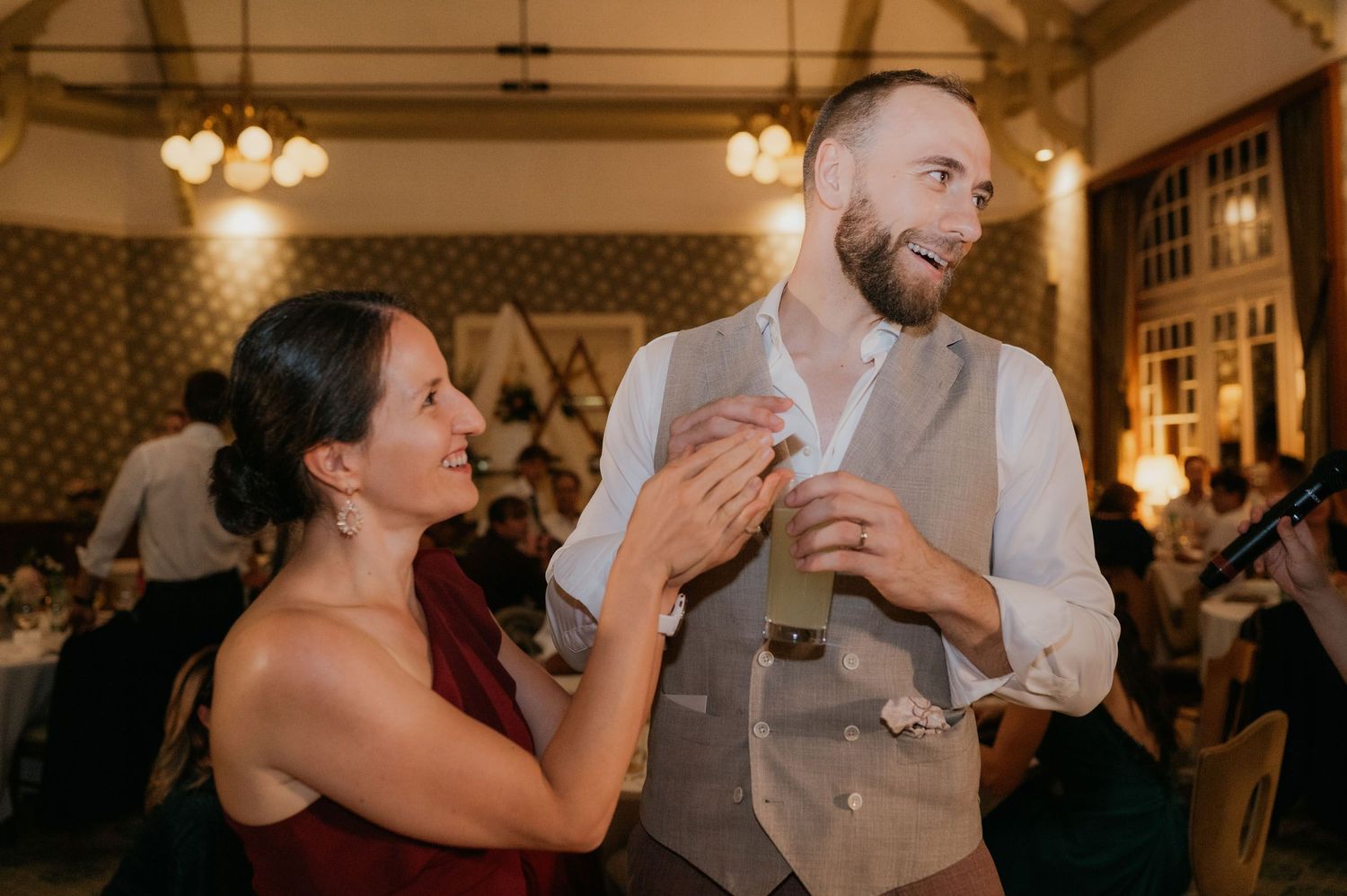 A smiling couple shares a joyful moment at a wedding reception, man in gray vest holding a drink.