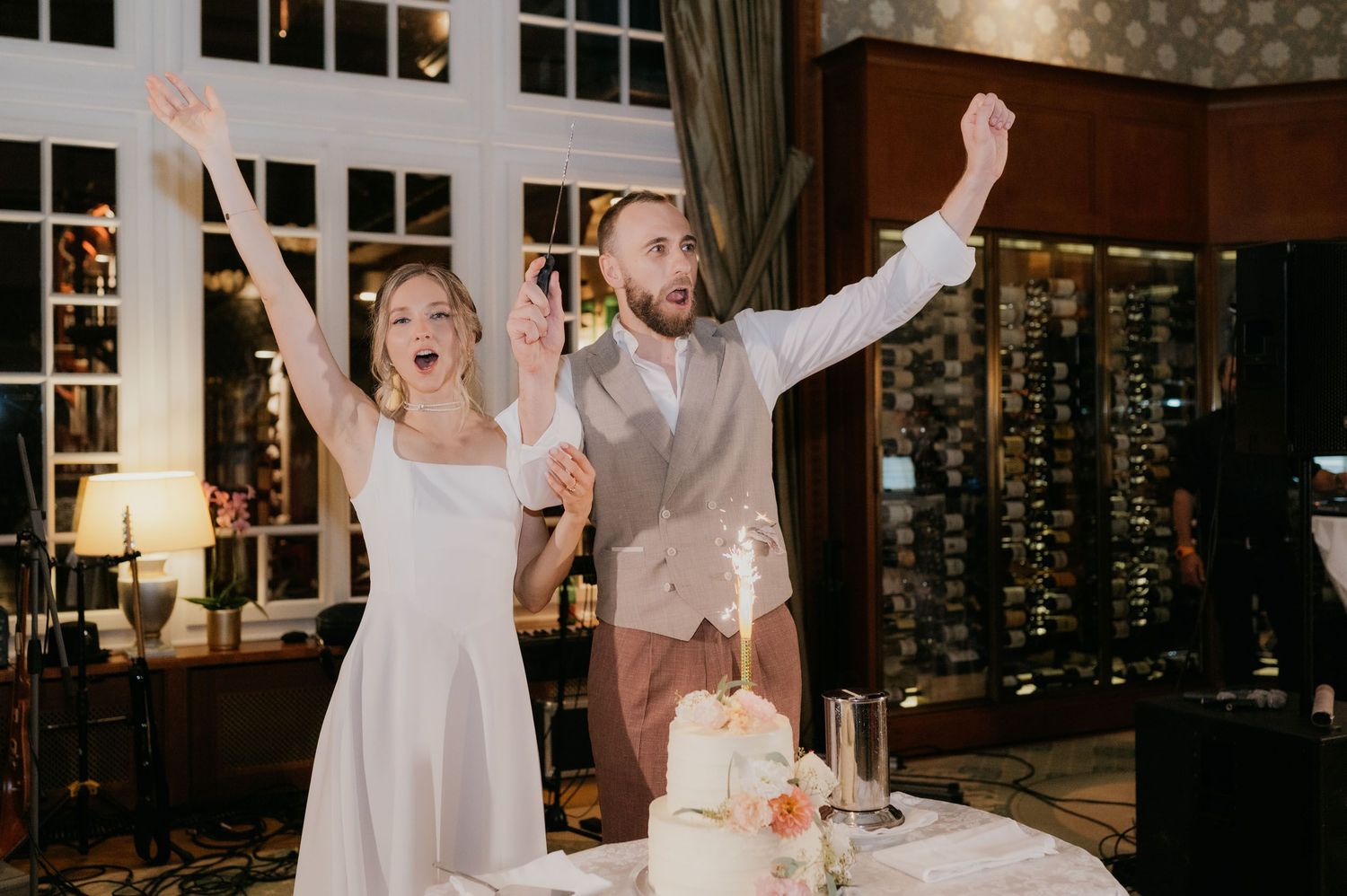 Bride and groom celebrating joyfully with raised arms after cutting their white wedding cake at an elegant reception venue.
