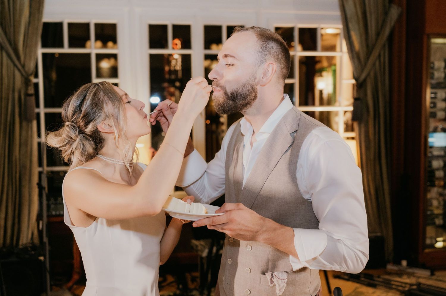 Bride feeding groom wedding cake at elegant indoor reception venue during cake cutting ceremony.