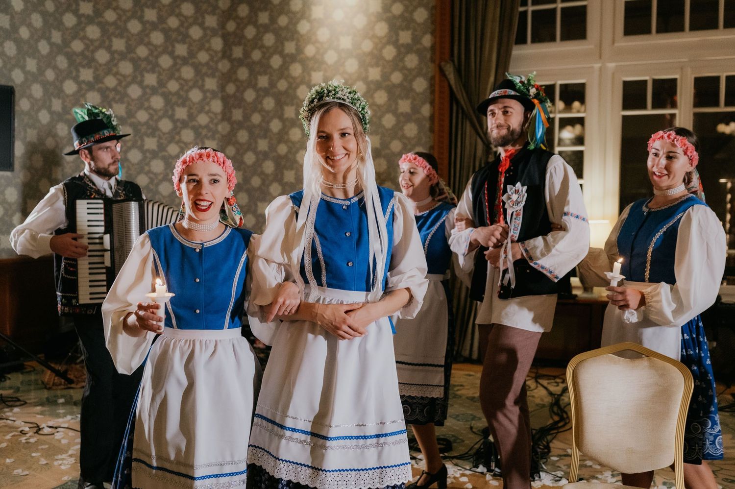 Traditional folk performers in blue and white costumes with floral wreaths playing accordion and clarinet indoors.
