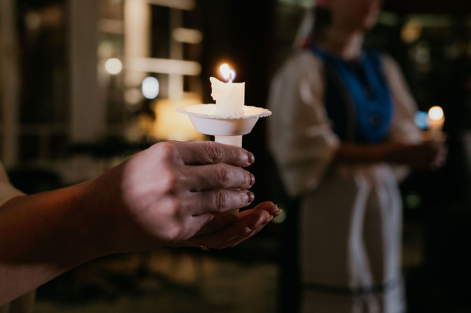 Hands holding a lit candle in a white holder during a candlelight vigil ceremony at night.
