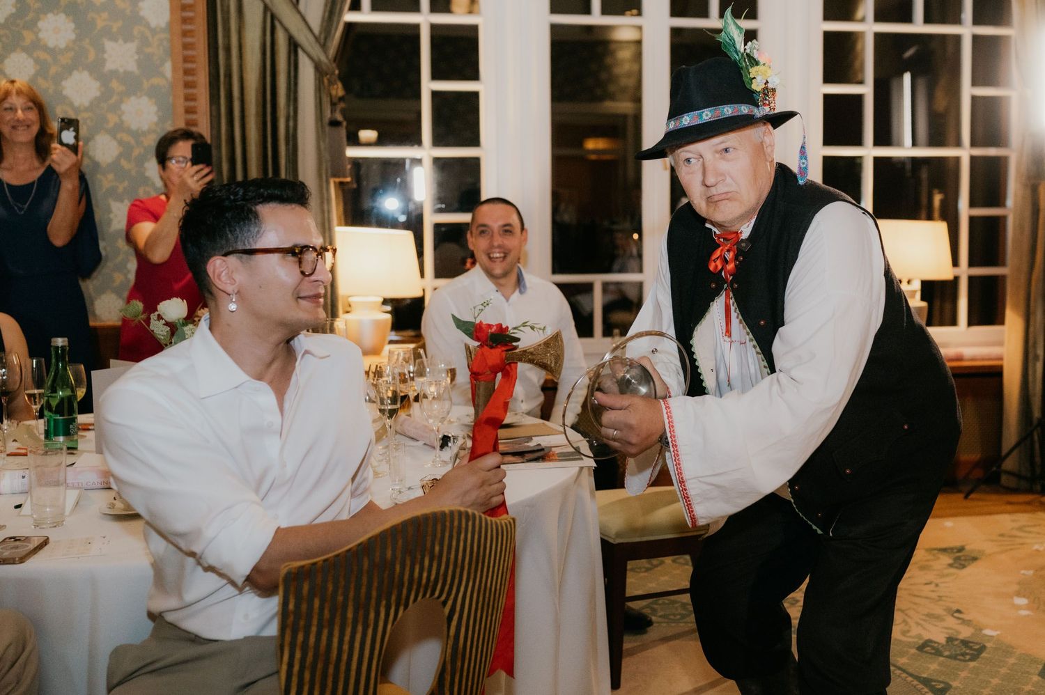 Man in traditional hat presents gift to seated guest at elegant indoor dinner party celebration.