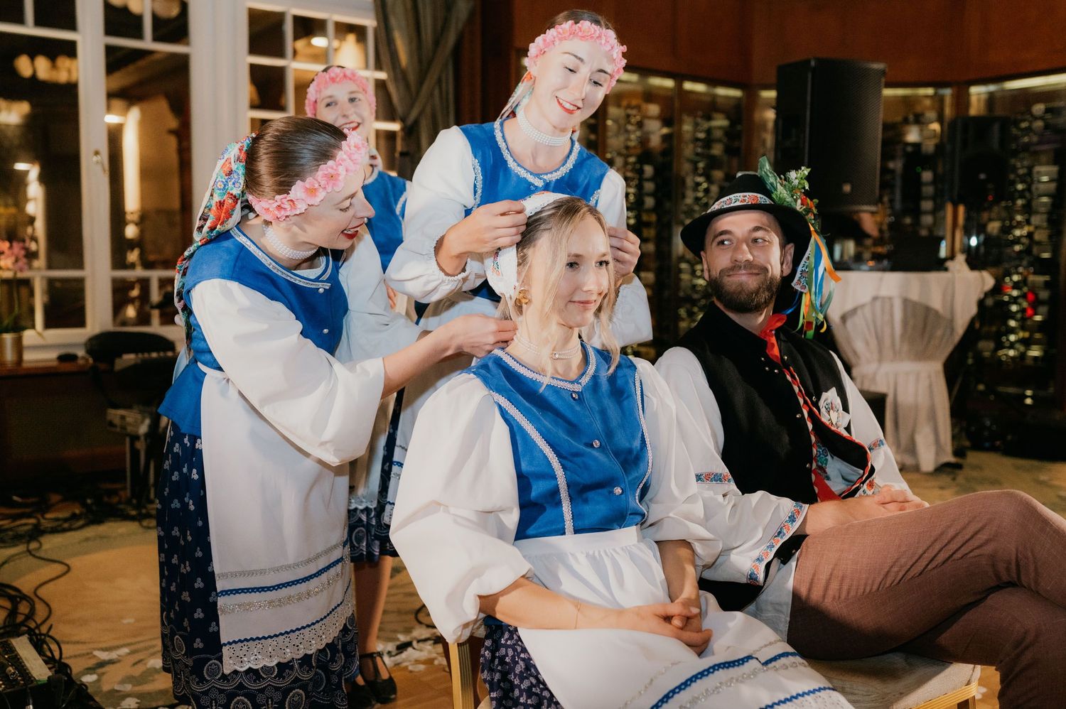 People in traditional Eastern European folk costumes braiding a womans hair at a festive indoor cultural event.