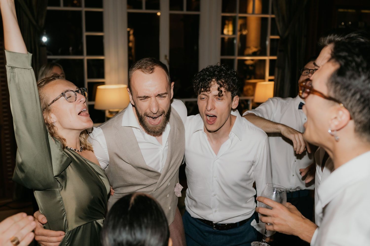 Group of friends celebrating joyfully at a wedding reception, singing and dancing together indoors.