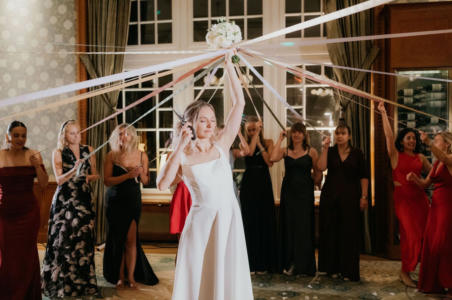 Bride tossing bouquet with ribbons at wedding reception while guests in formal attire watch and cheer.