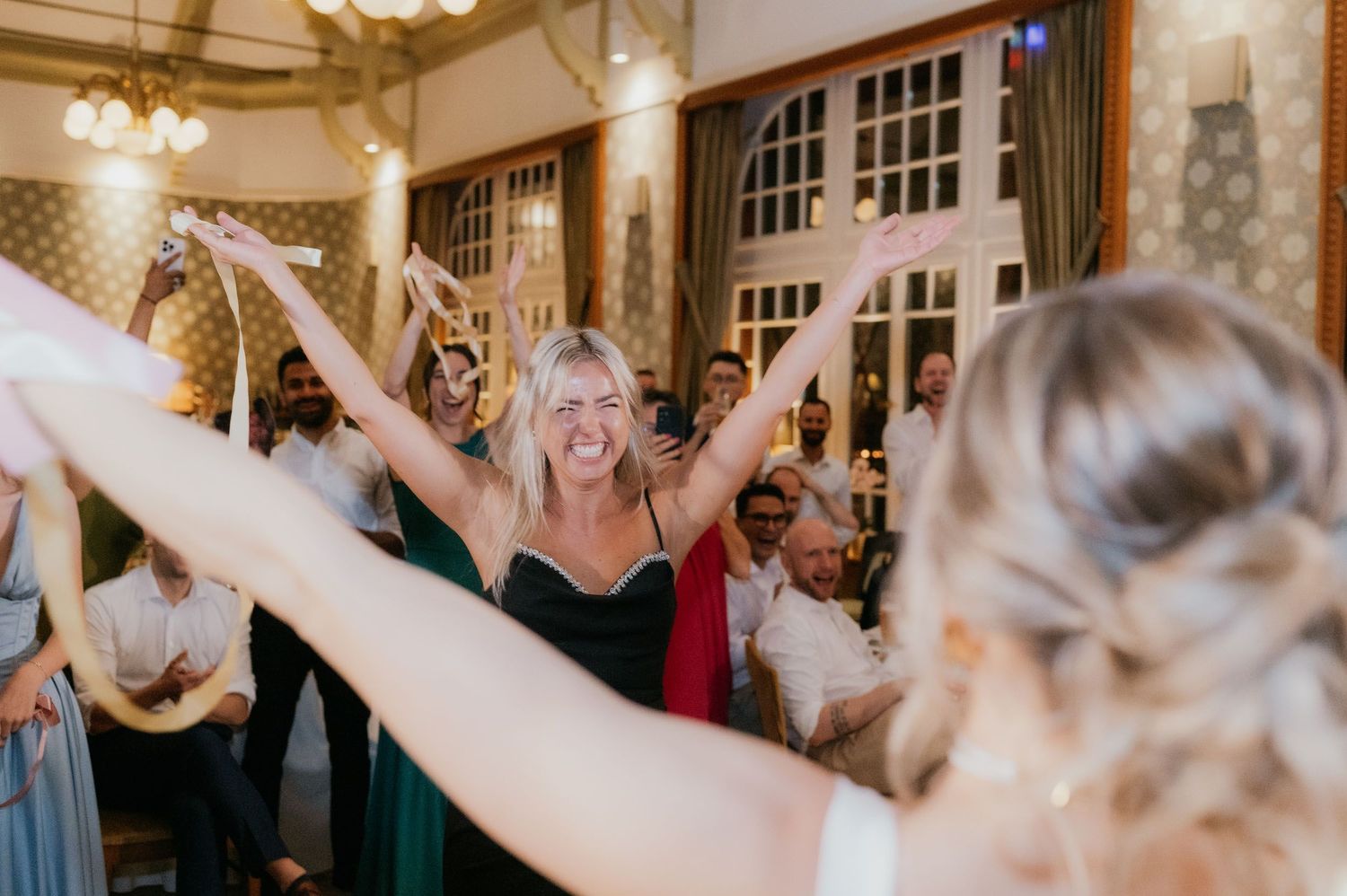 Joyful wedding guest in green dress raises arms celebrating on dance floor at elegant indoor reception.