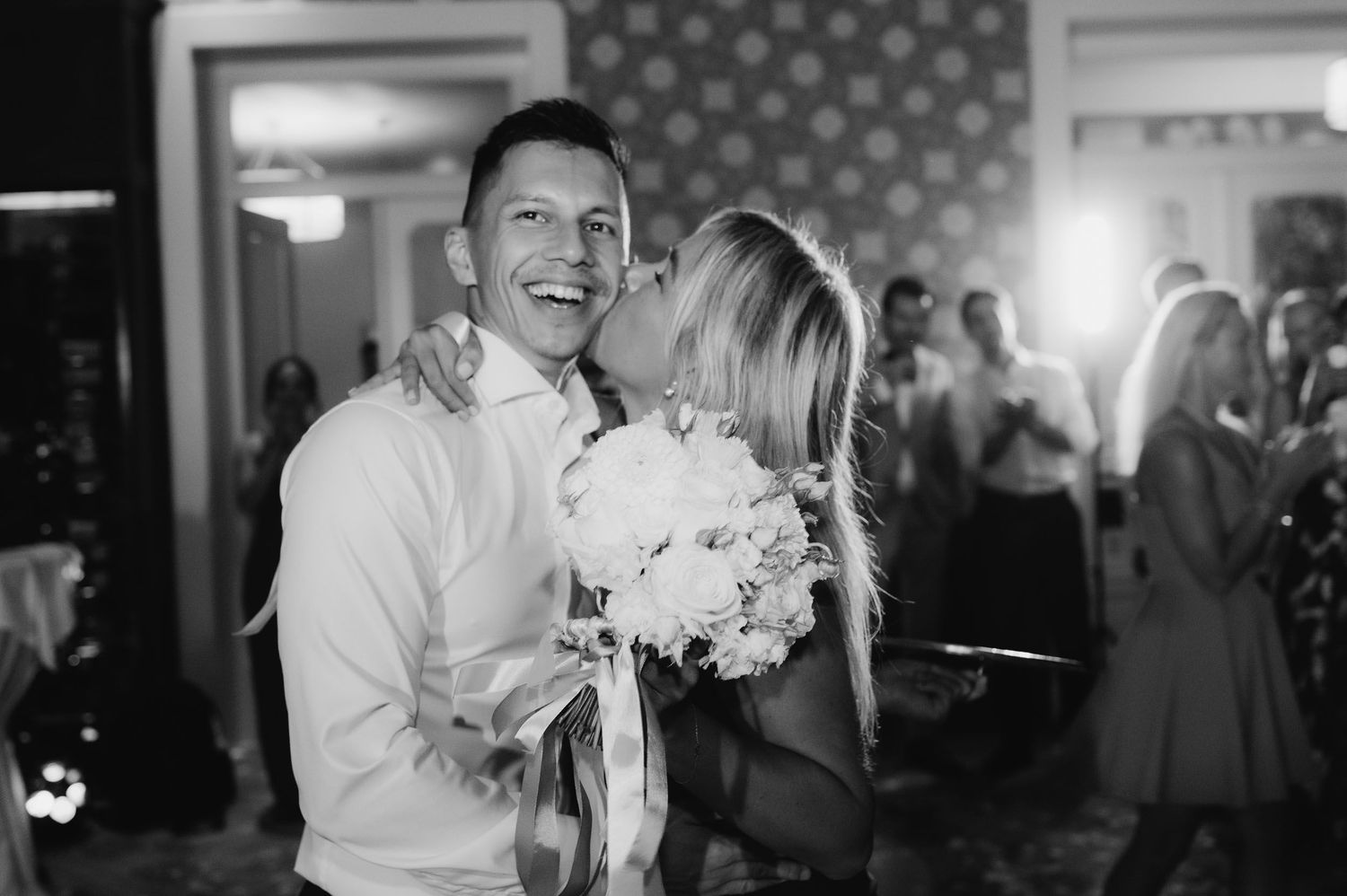 Happy groom laughs as bride kisses his cheek holding white bouquet at wedding reception.