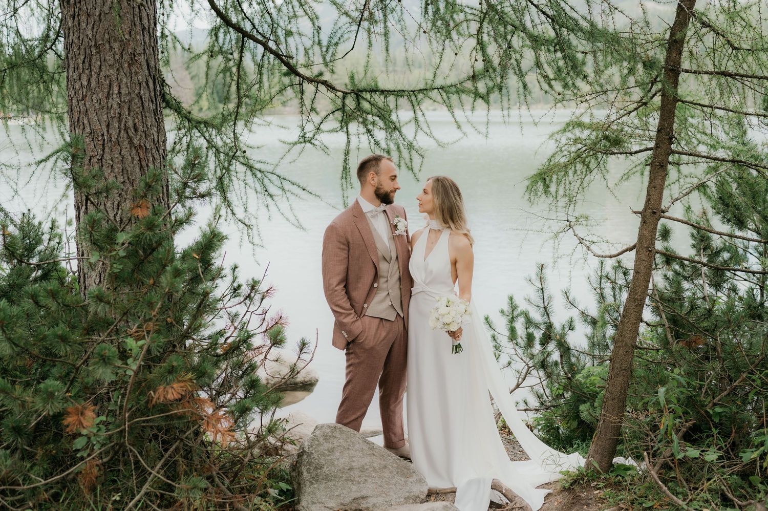 Bride and groom stand among pine trees by a misty lake, bride in white gown, groom in tan suit.