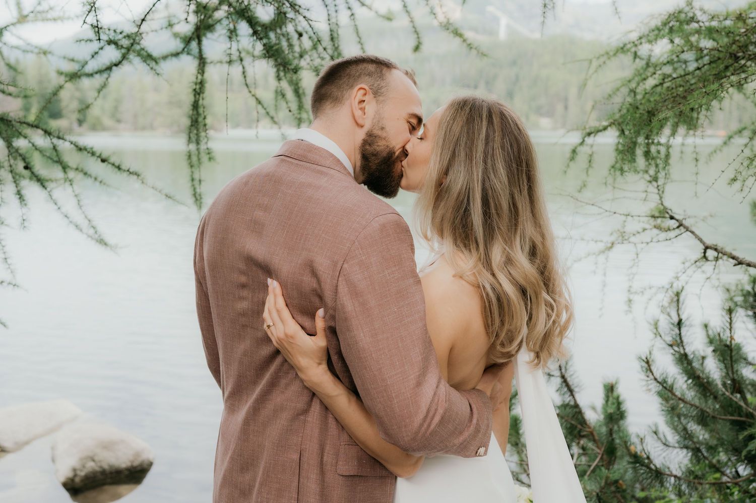 A couple shares a romantic kiss outdoors near a serene lake, surrounded by lush green pine trees.