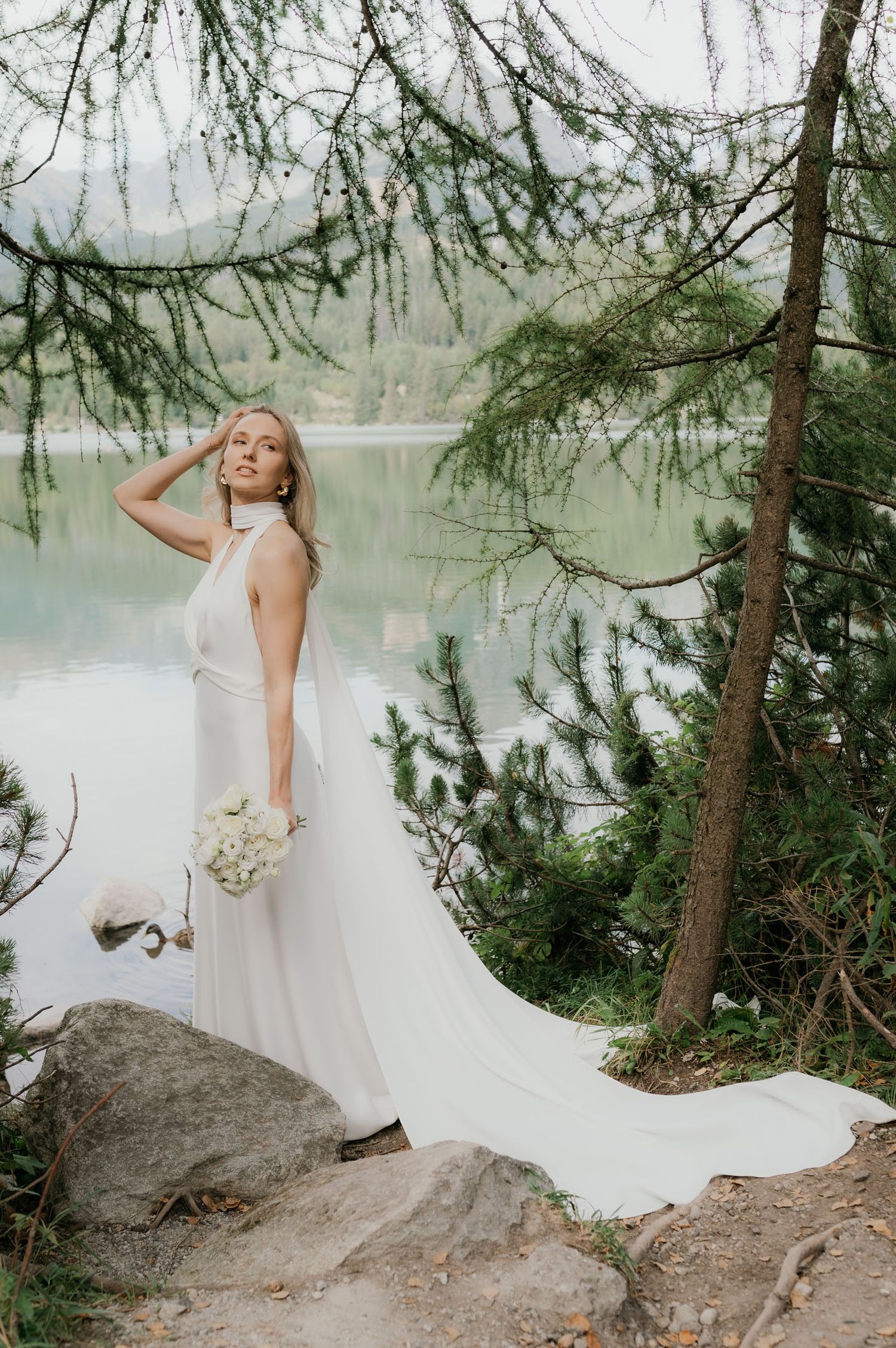 Bride in white halter gown with long train holds bouquet by a serene lake surrounded by pine trees.