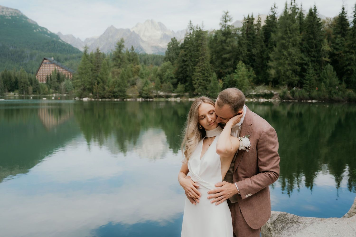 Couple embracing by a mountain lake with forest and peaks reflected in calm water on their wedding day.