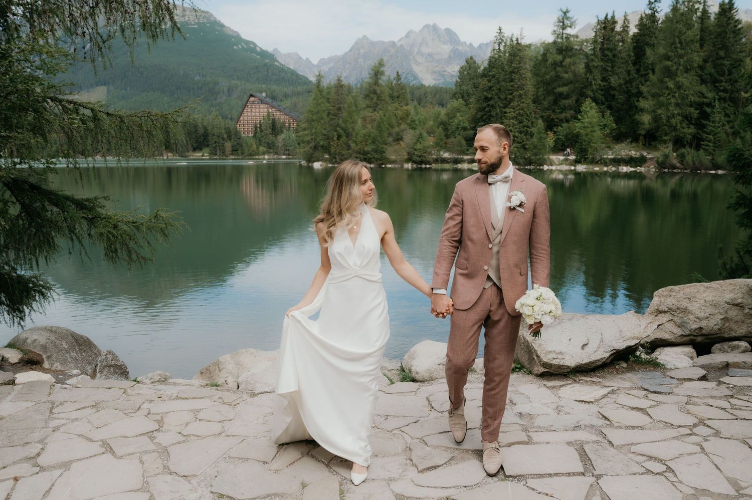Bride and groom holding hands by a mountain lake surrounded by pine trees and peaks on their wedding day.