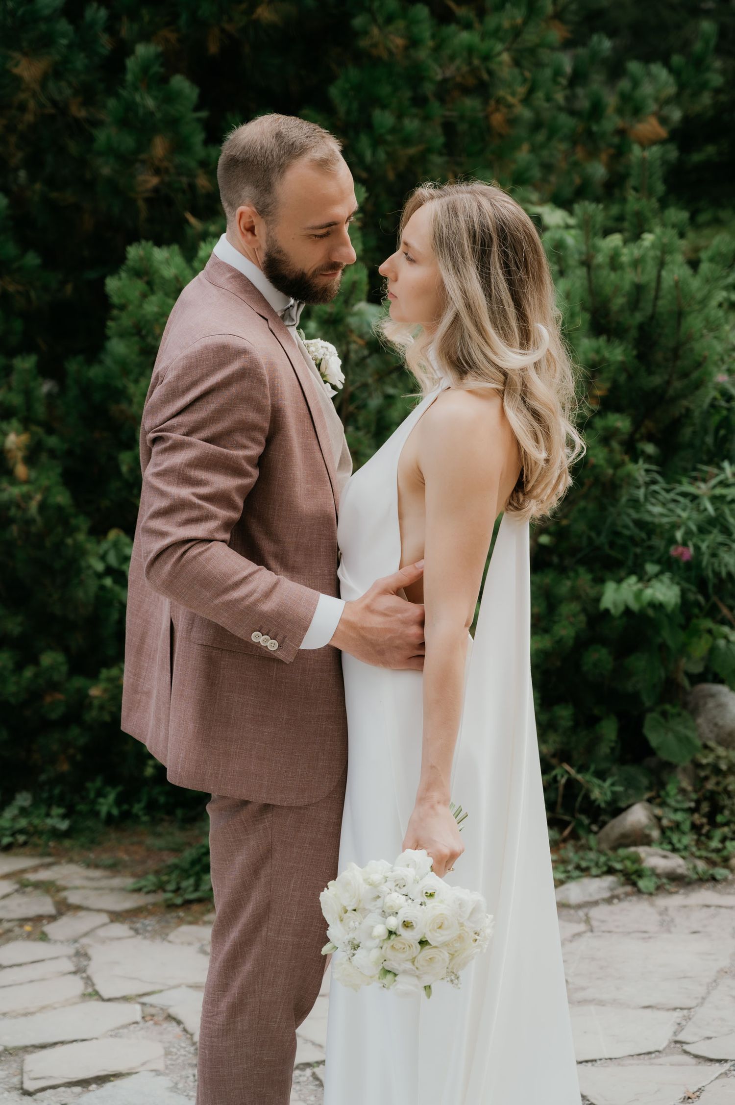 A bride and groom share a romantic moment outdoors, she holds white flowers in a white gown, he wears a dusty rose suit.