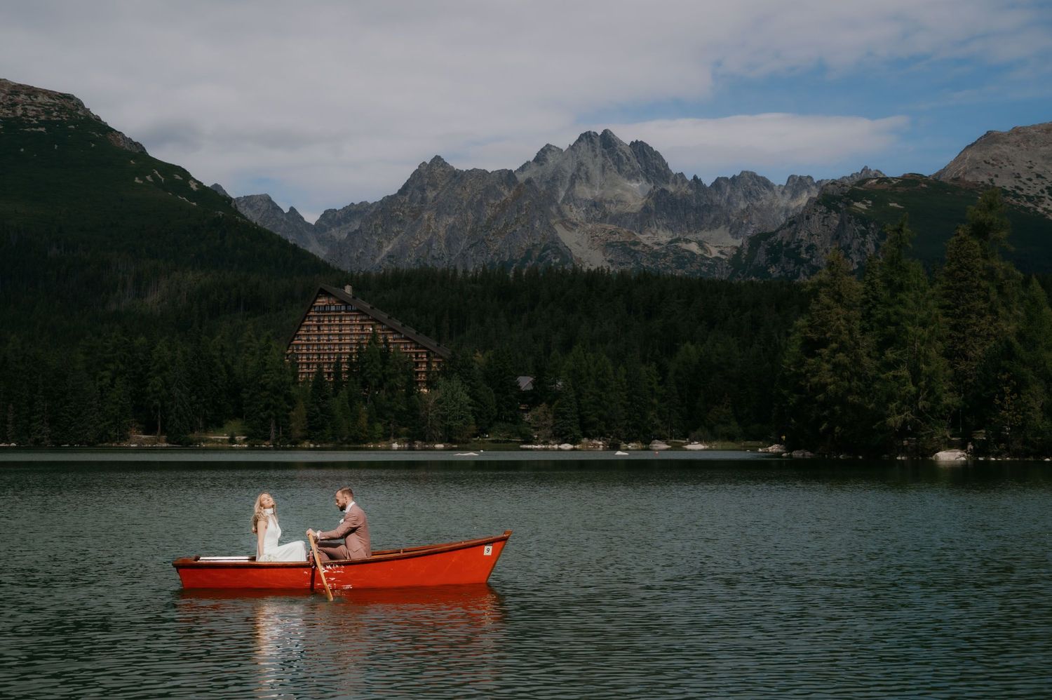 Couple in a red rowboat on a mountain lake with dense forest and dramatic peaks in the background.