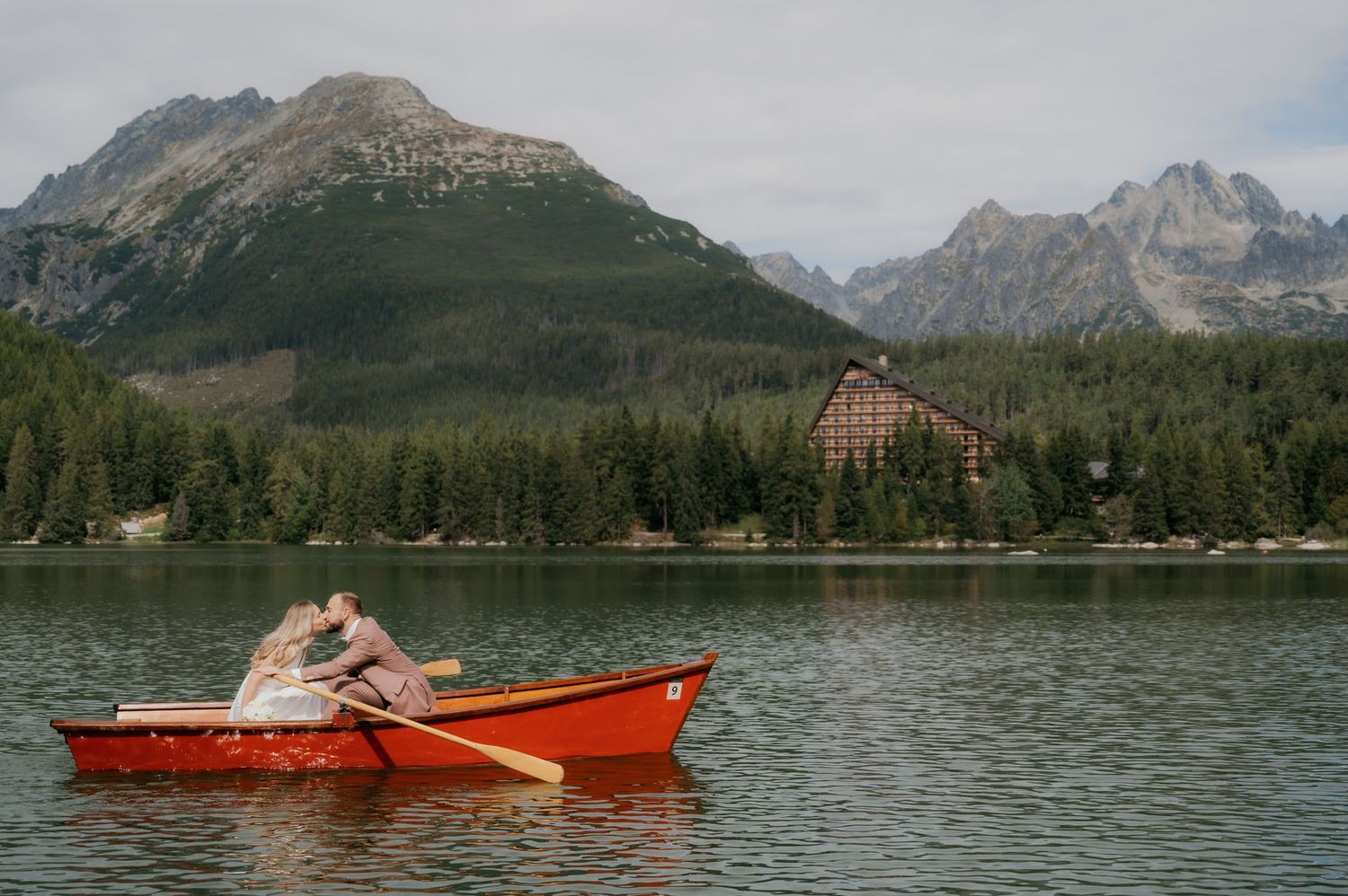 Couple embracing in a red rowboat on a mountain lake with forested slopes and a chalet in the background.