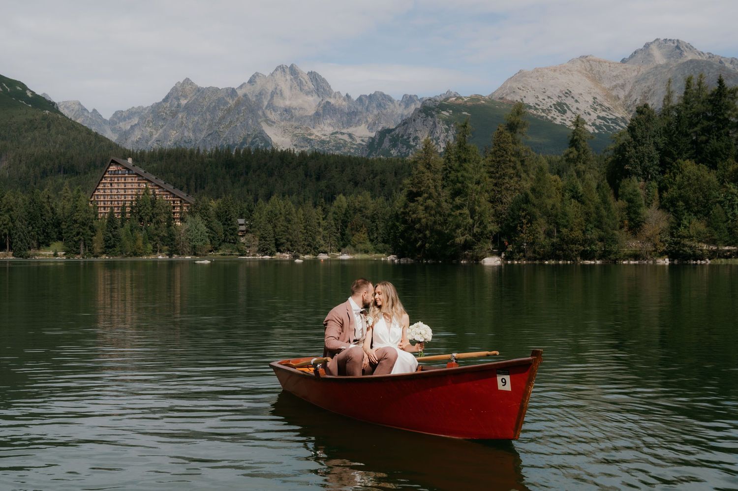 Couple sharing a romantic kiss in a red rowboat on a serene mountain lake with forest and peaks behind.
