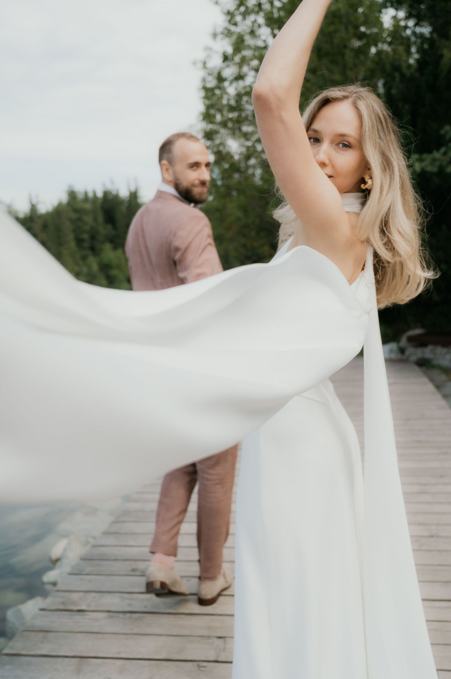 A bride in a flowing white dress twirls on a wooden dock while her groom stands smiling in the background.