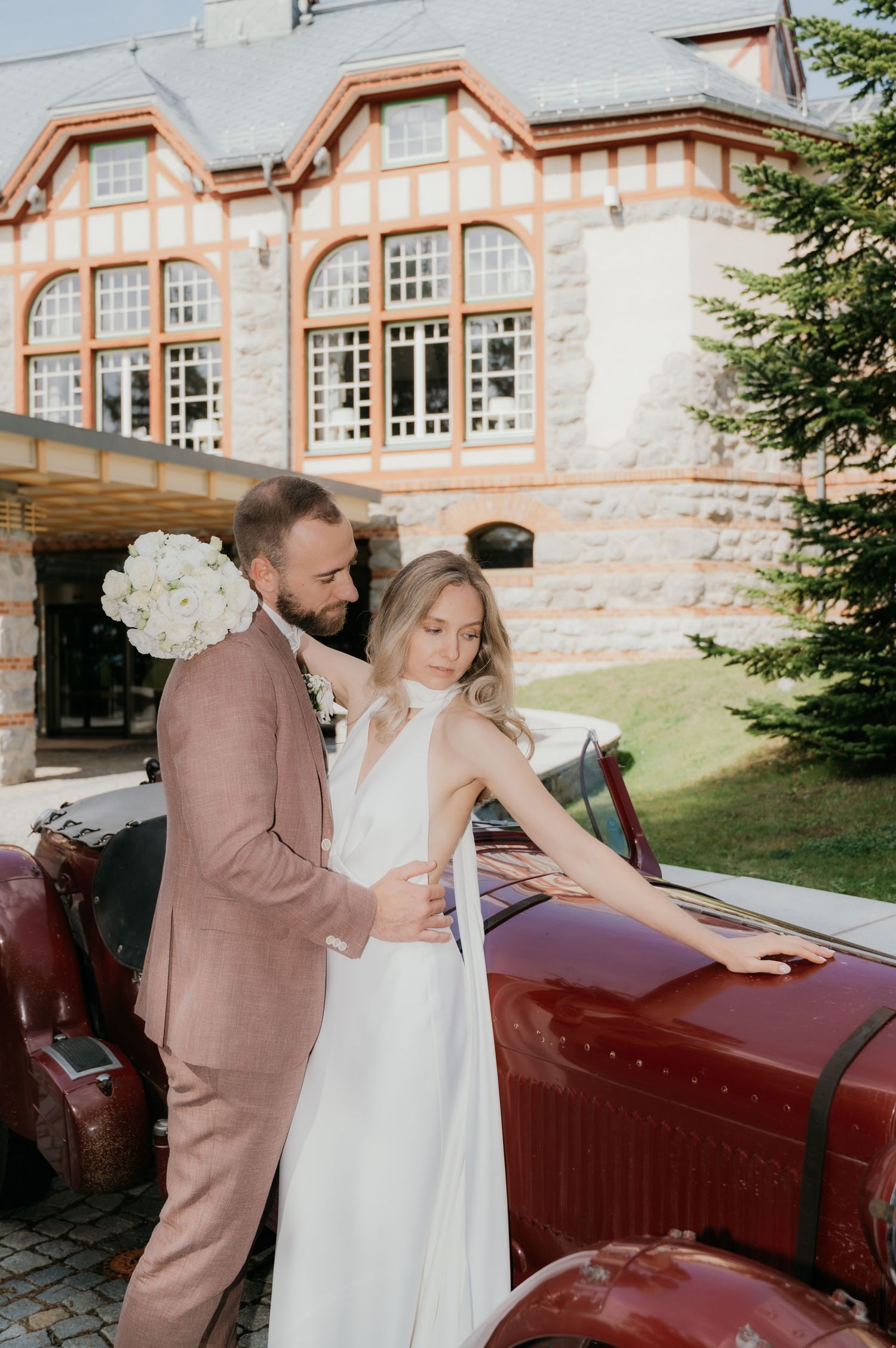 Bride in white gown and groom in pink suit pose beside vintage red car outside elegant European manor.