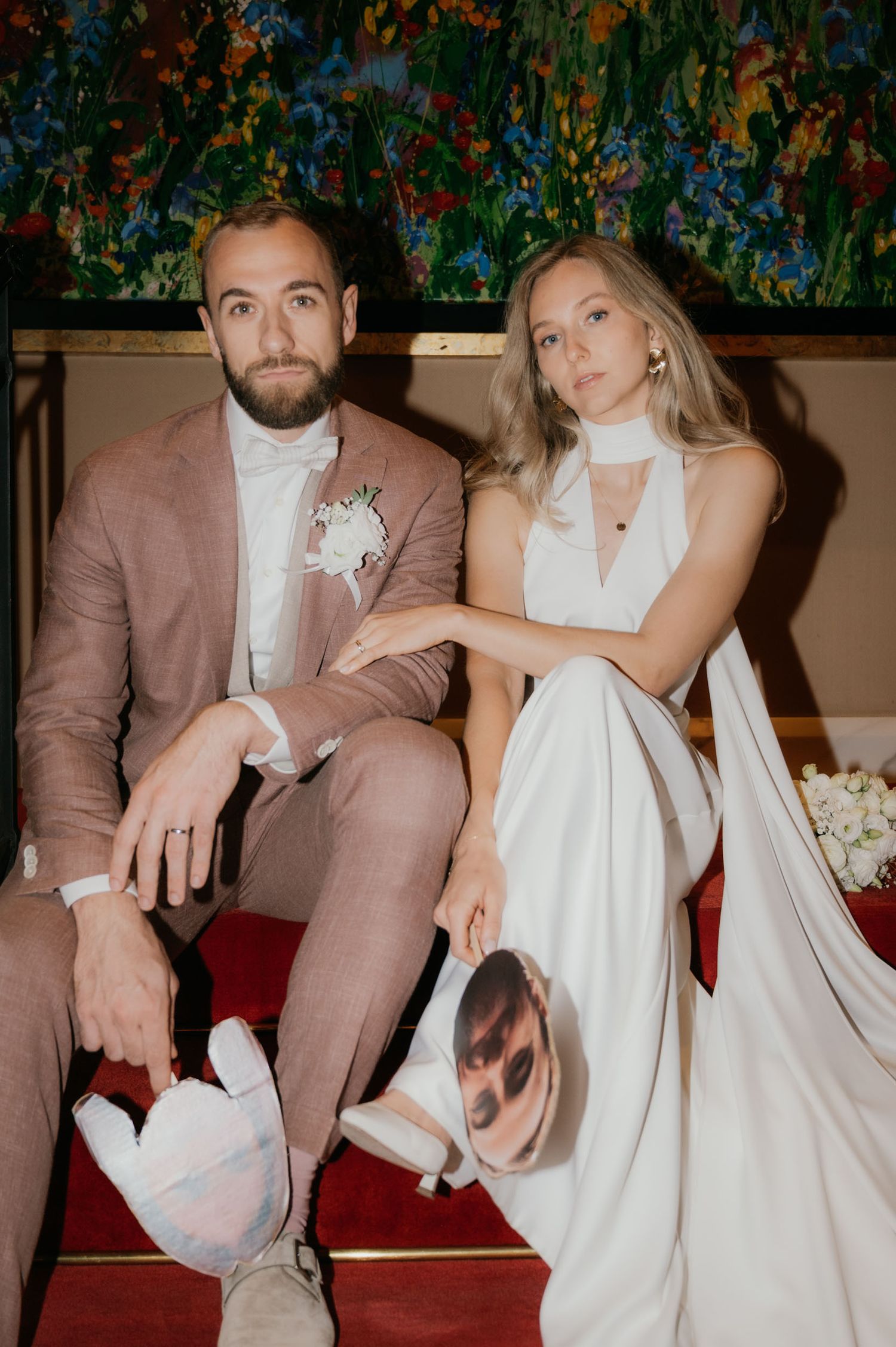 Bride in white halter gown and groom in dusty rose suit sit together holding decorative pillows before colorful floral painting.
