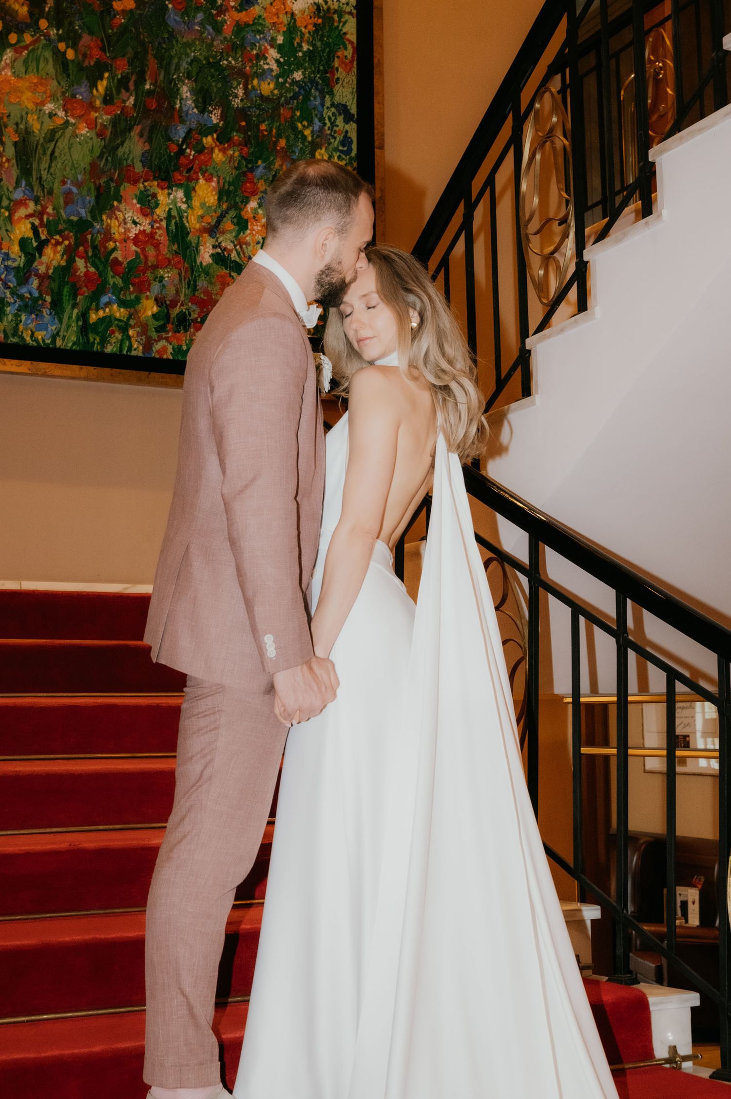 Bride in backless white gown and groom in blush suit hold hands by red carpeted staircase near colorful painting.