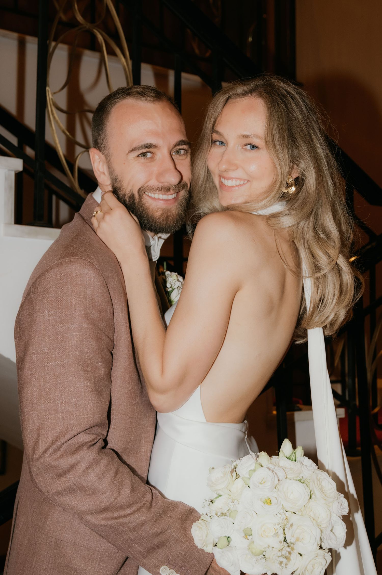 A smiling bride in a backless white gown holds white flowers while embracing a bearded groom in a mauve suit.