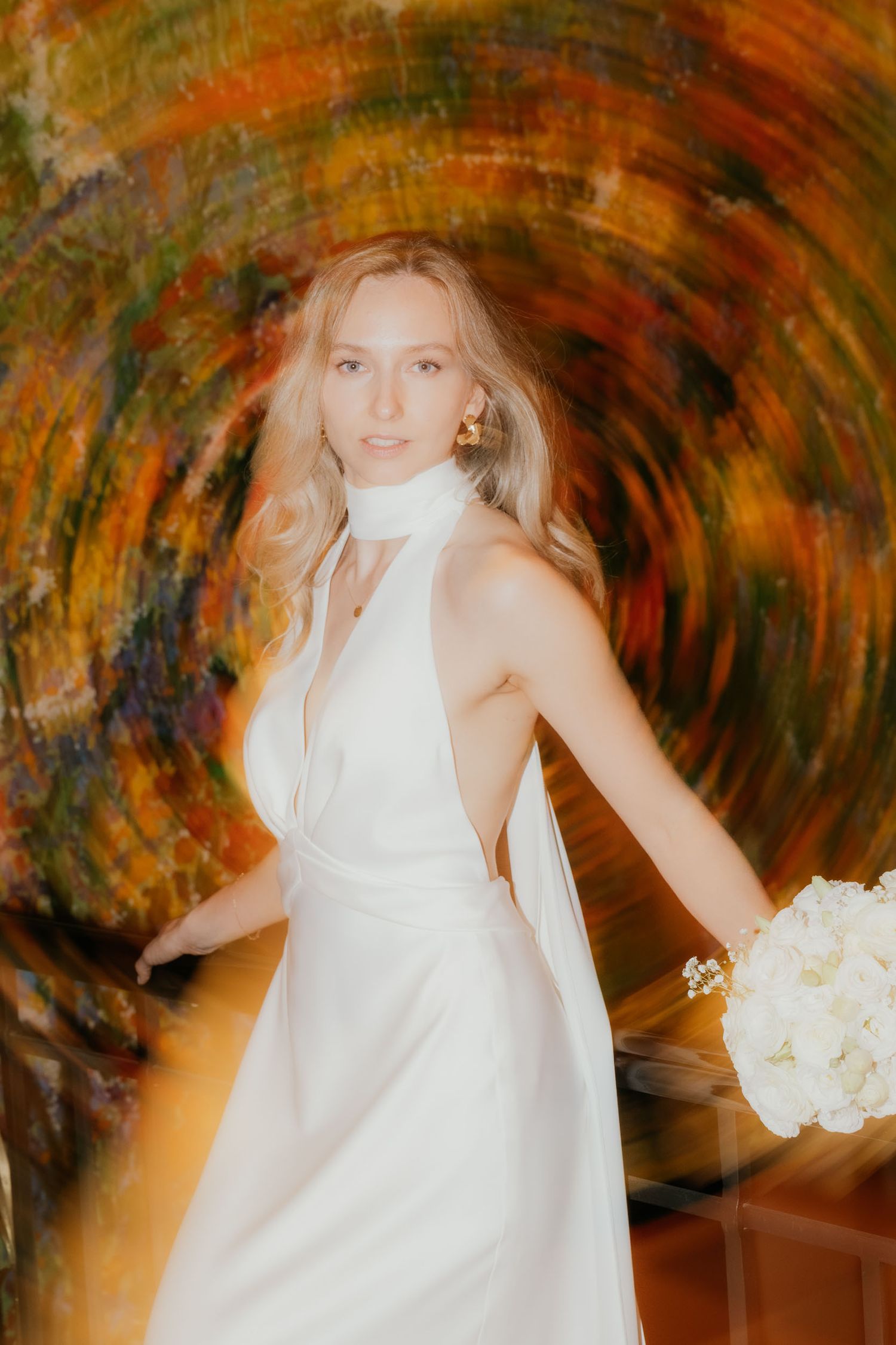 Bride in white halter gown holding bouquet, framed by swirling colorful abstract background in artistic wedding portrait.