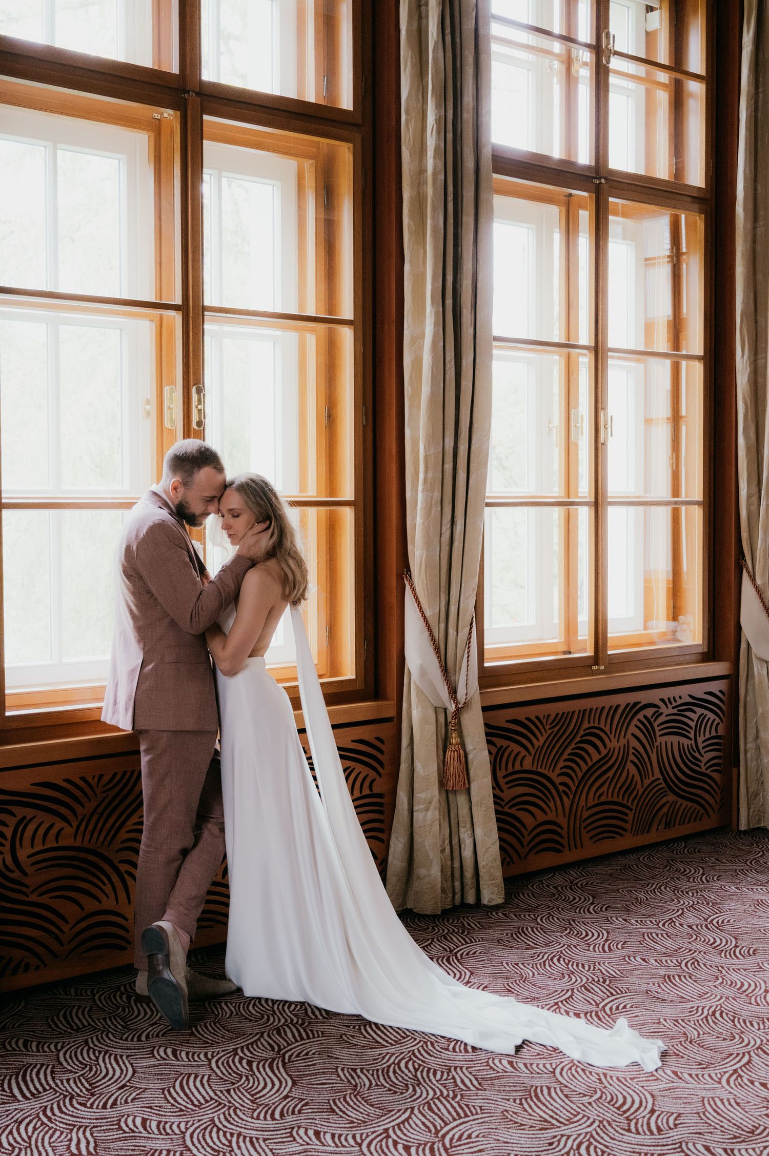 Bride and groom embracing by large wooden windows in elegant venue with patterned carpet and drapes.