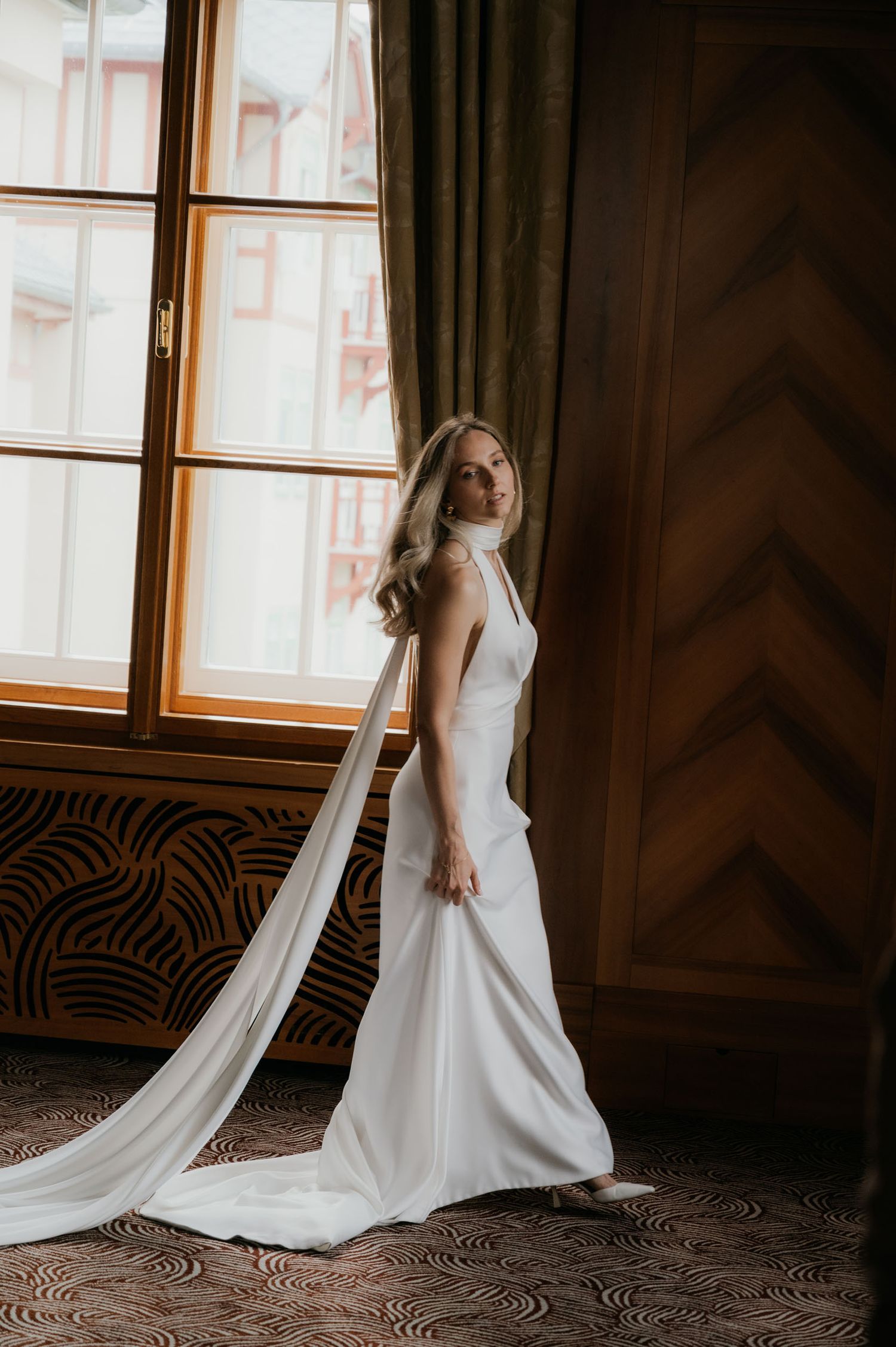 Bride in elegant white halter wedding gown with long train stands by large window in ornate room.