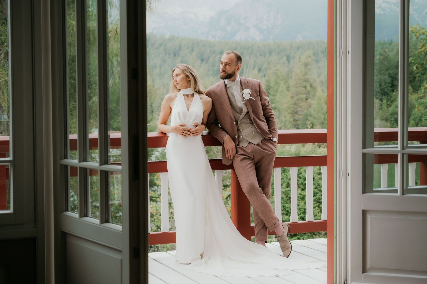 Bride in white gown and groom in brown suit pose on a wooden balcony with mountain forest views.