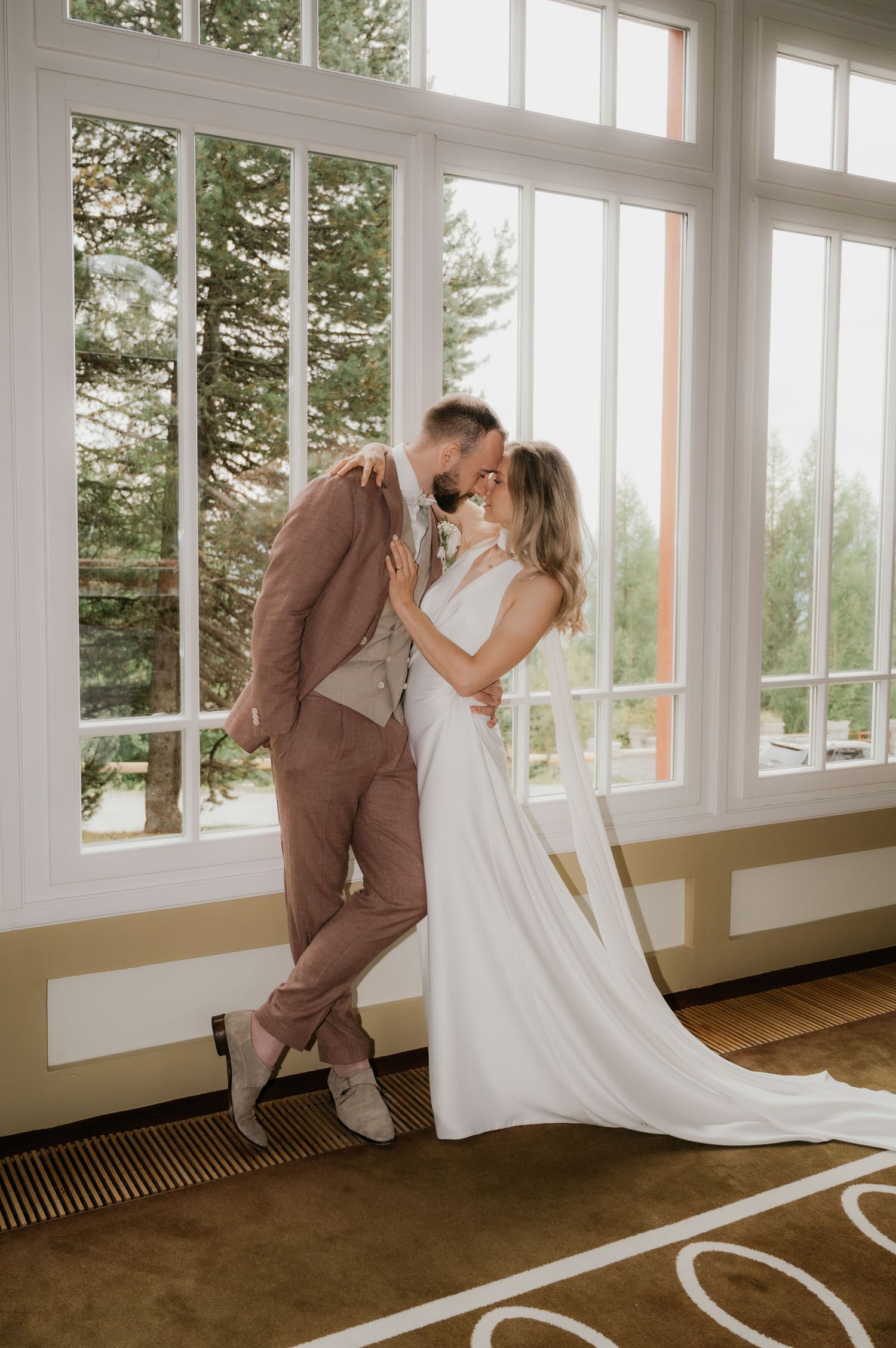 Bride and groom sharing a romantic kiss in front of large windows, bride in white gown, groom in dusty rose suit.