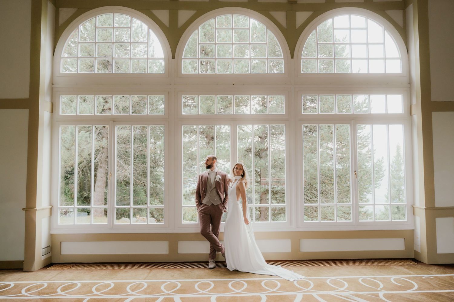 Bride and groom share a romantic moment in front of large arched windows in an elegant wedding venue ballroom.