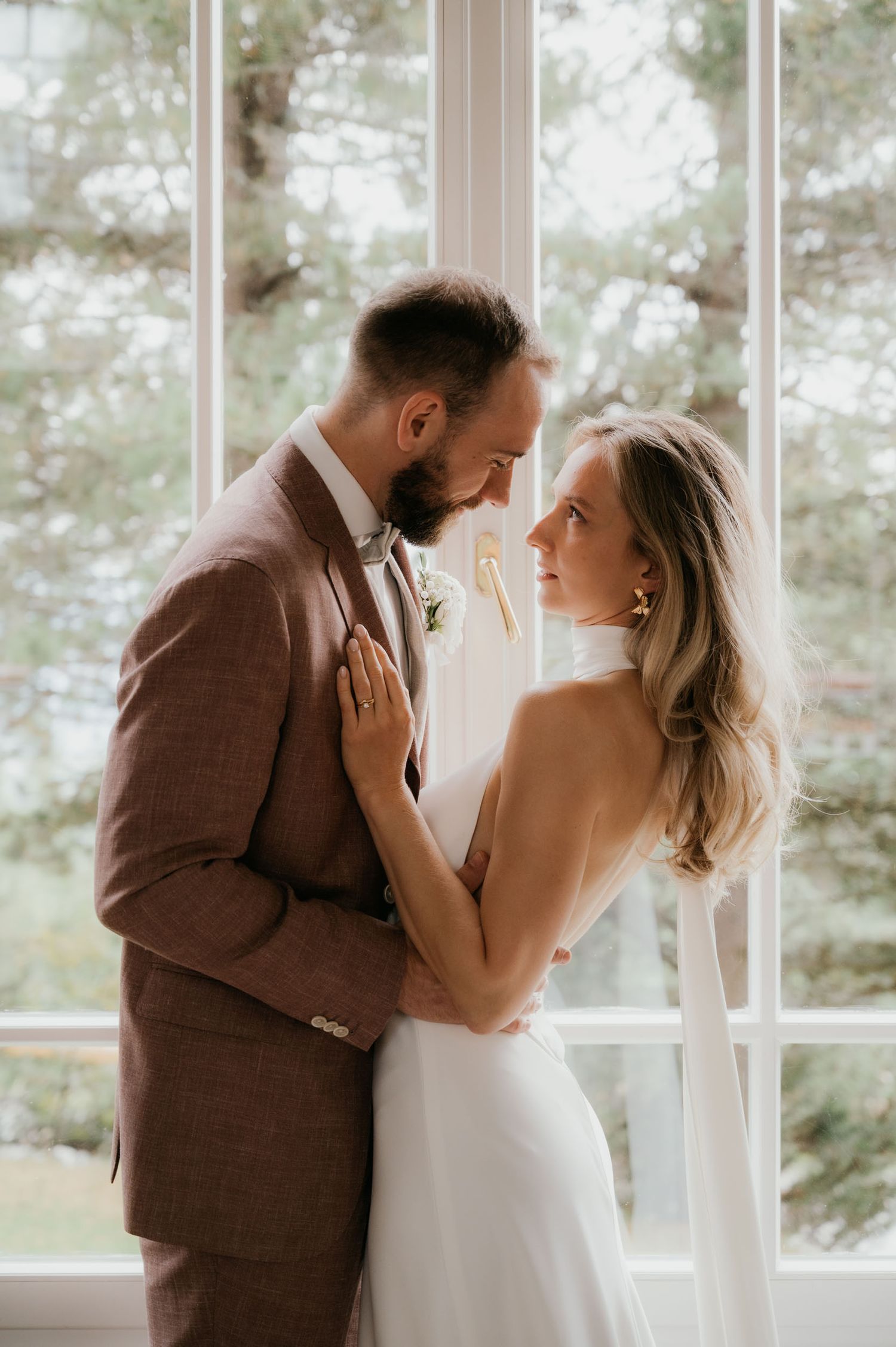 A bride and groom share an intimate moment by a sunlit window, she in a white gown, he in a brown suit.