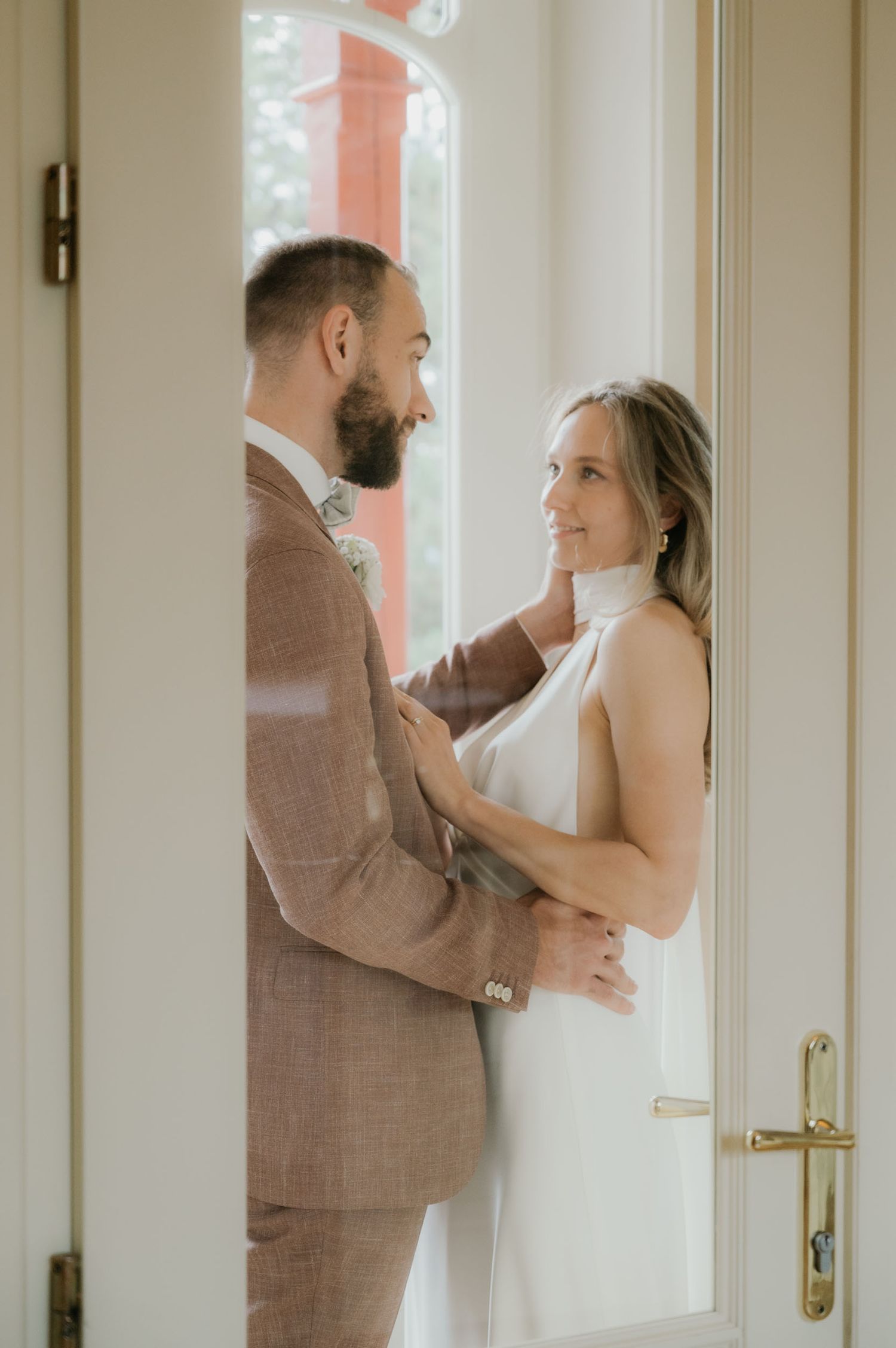 A couple shares a tender moment in a doorway, the groom in a tan suit and bride in a white halter dress.