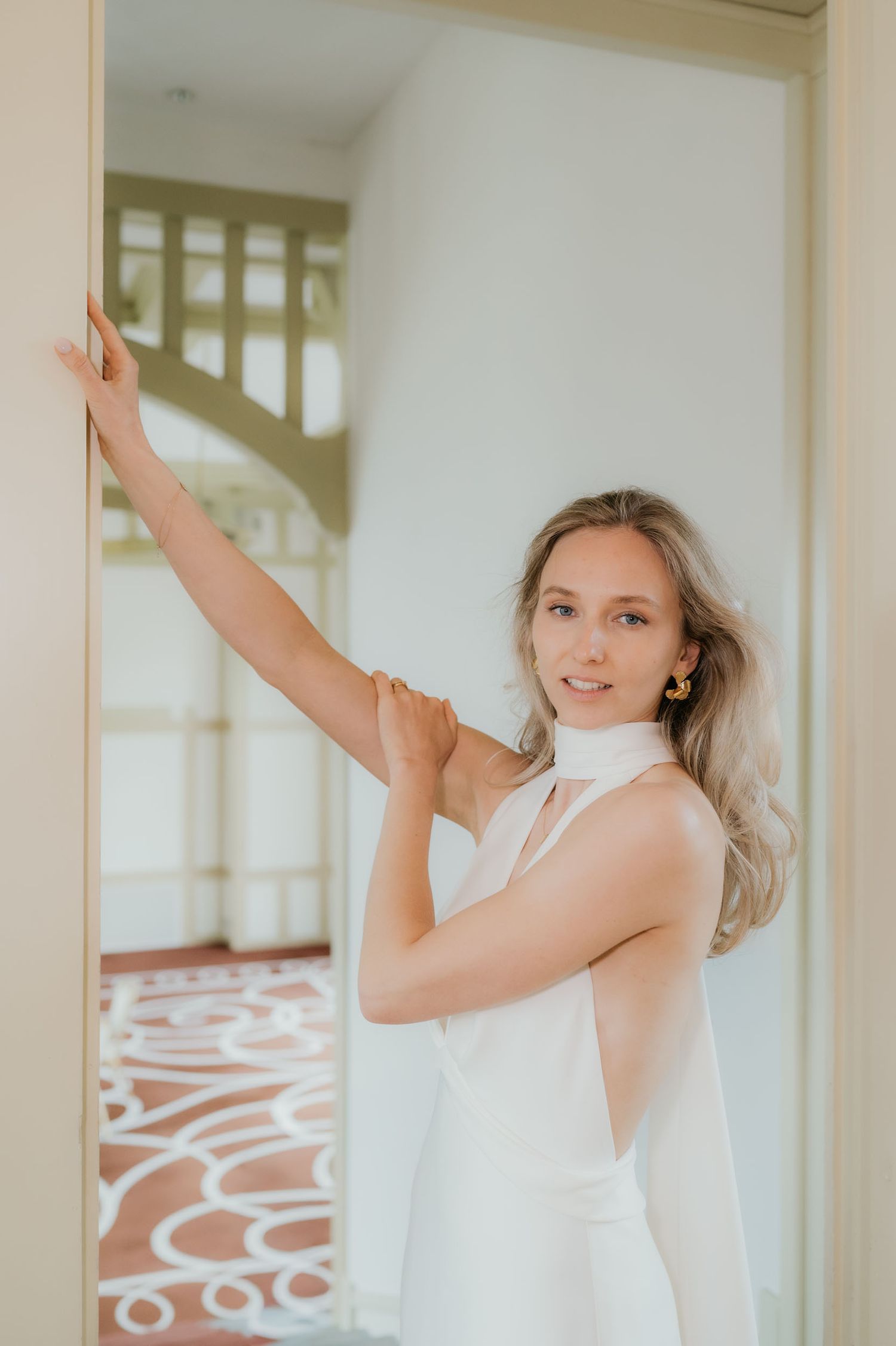 A young woman in a white halter dress poses gracefully in a doorway, with a spiral staircase visible behind her.