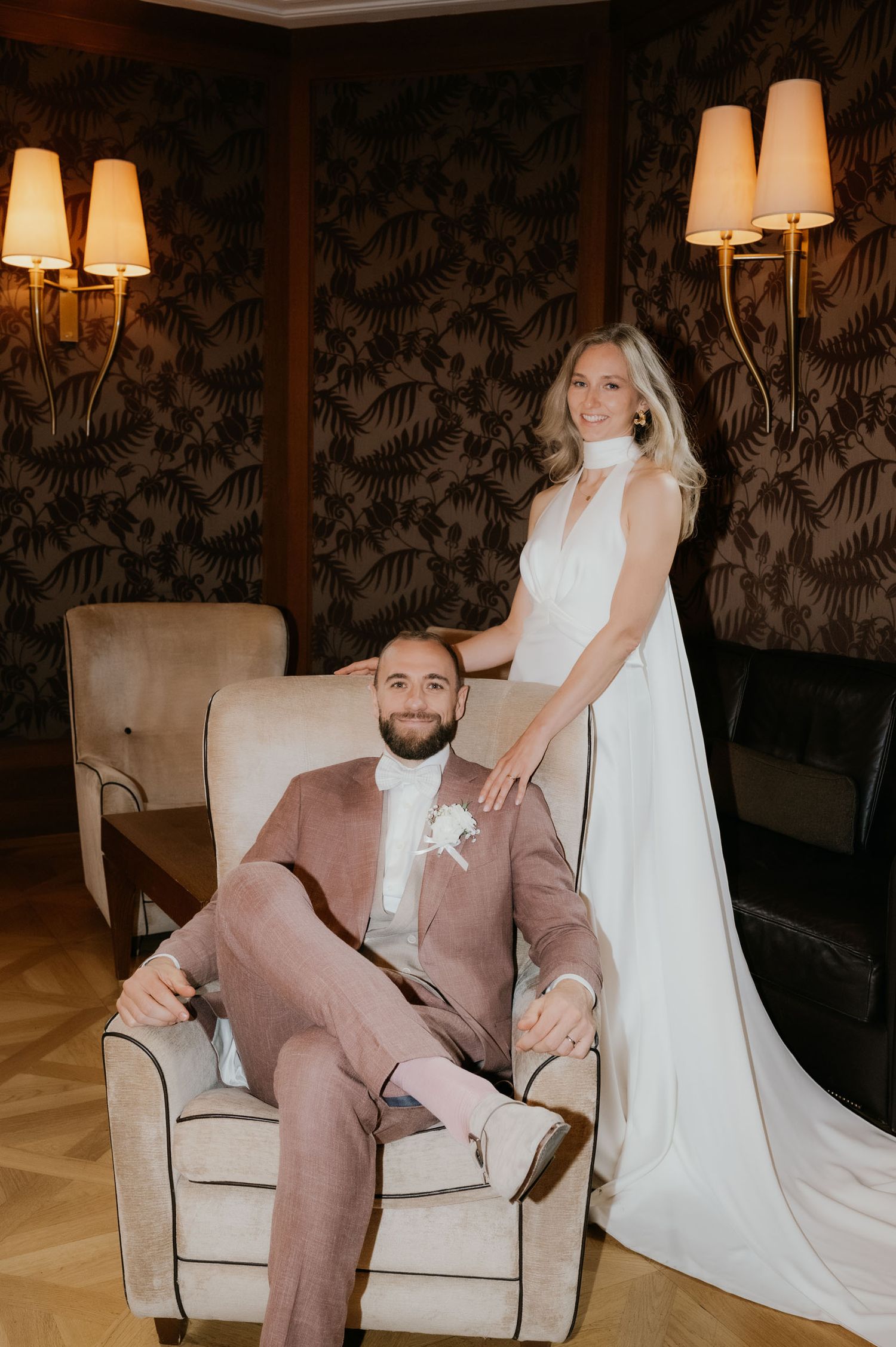 Bride in white gown stands behind groom in pink suit seated in armchair, posing in elegant room with floral wallpaper.
