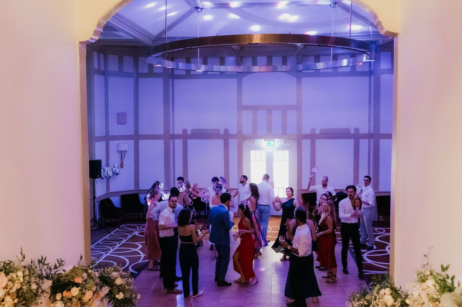 Elegant wedding reception with guests dancing in a grand ballroom with arched ceilings and soft purple uplighting.