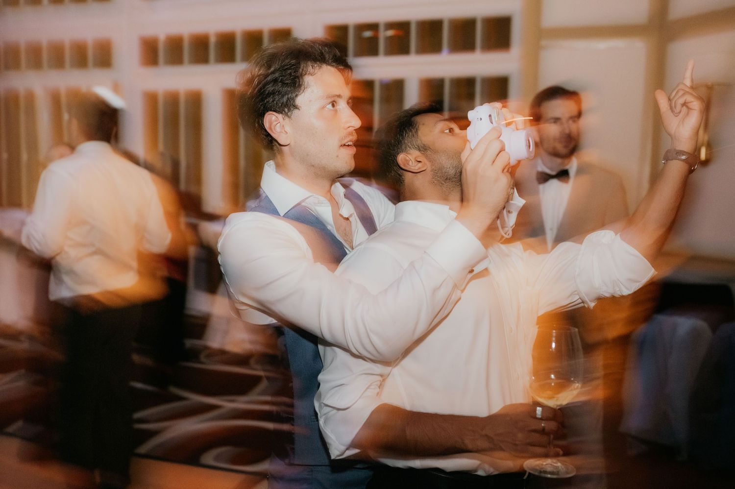 Two grooms share a joyful dance moment at their wedding reception, one lifting the other while guests celebrate.