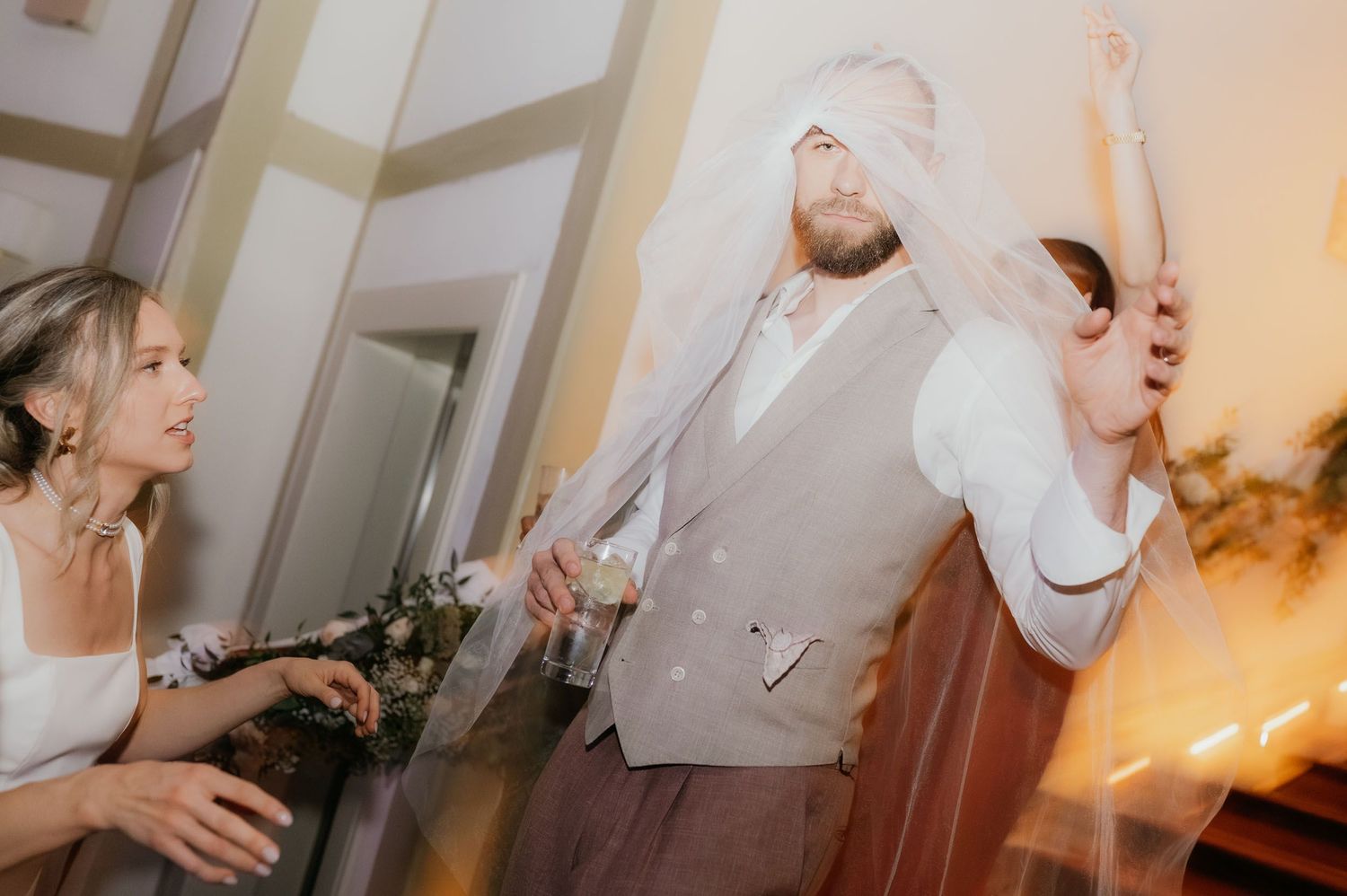 A groom in a vest laughs as a bride playfully drapes her wedding veil over his face at a reception.