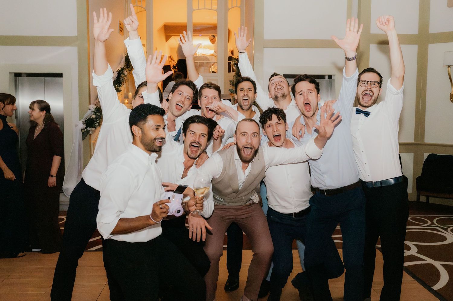 Excited group of men celebrating at a wedding reception, raising hands and smiling in a elegant ballroom.