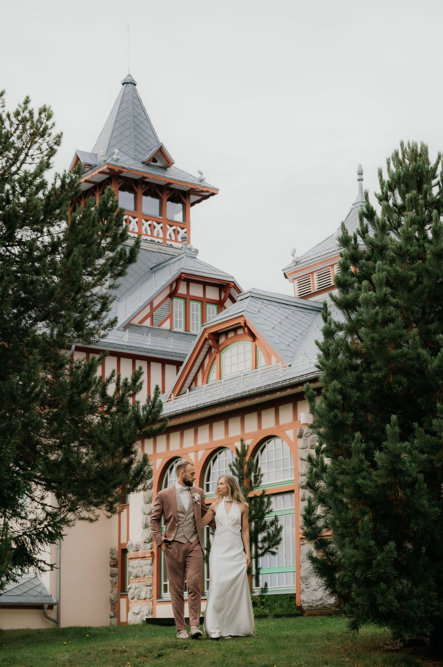 Bride and groom standing together in front of a charming Victorian-style building surrounded by pine trees.