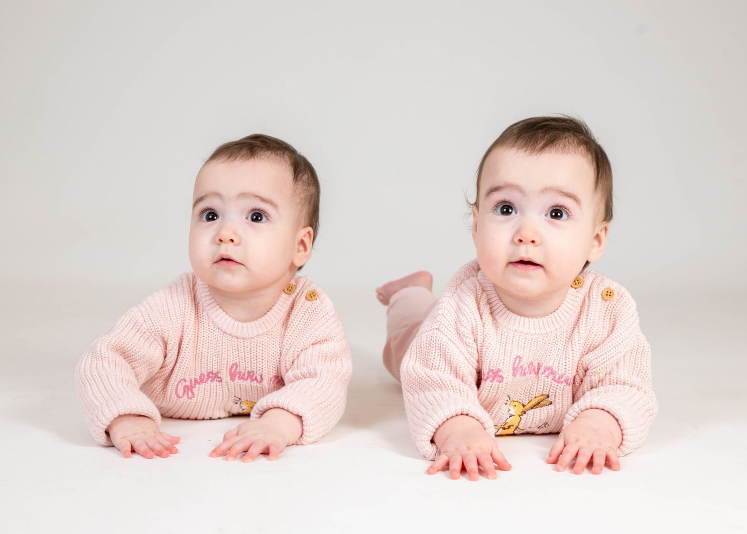 Twin babies in matching pink outfits laying on their tummies on a white background.