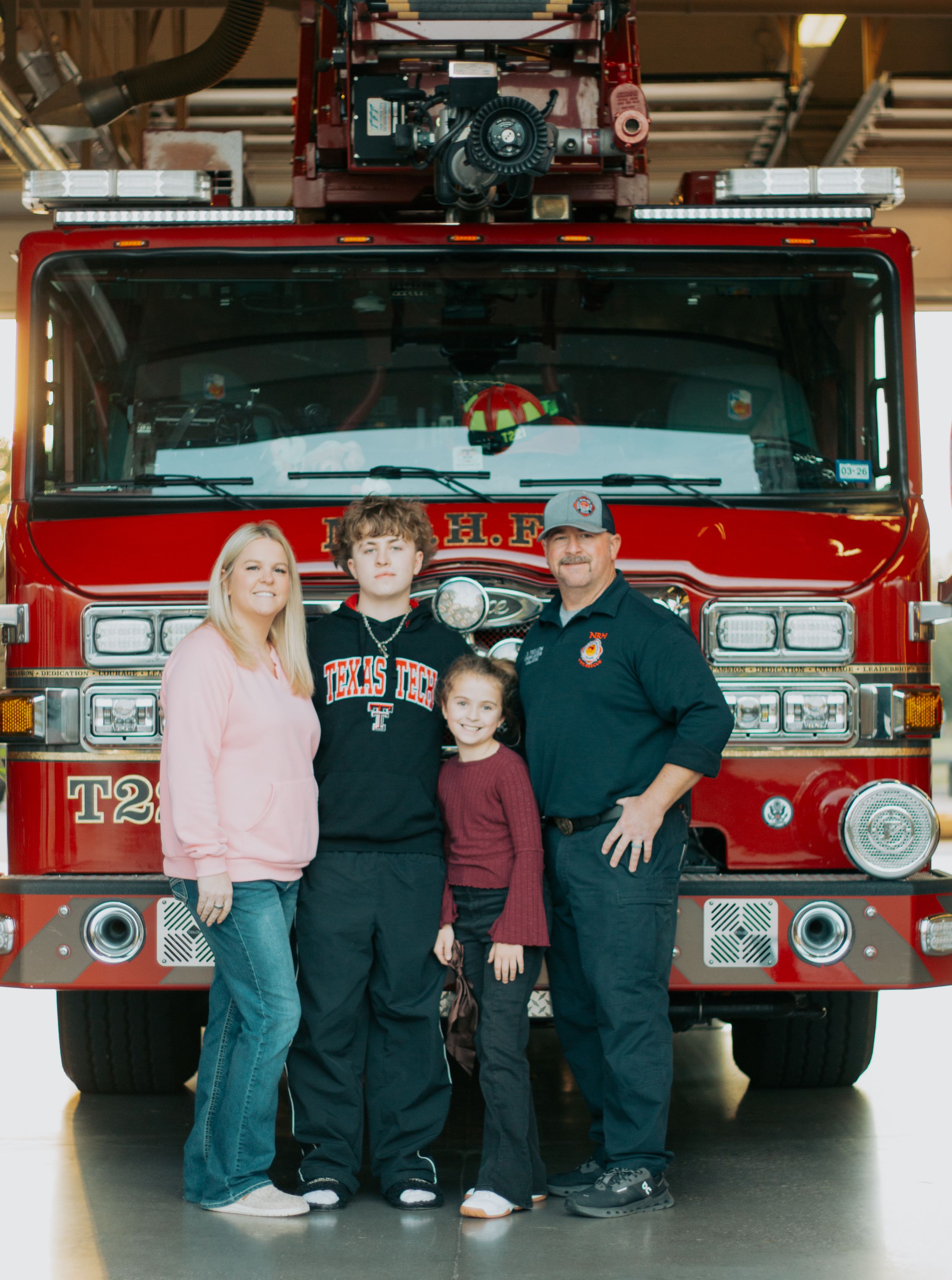 Four firefighters pose in front of red fire trucks at a fire station.