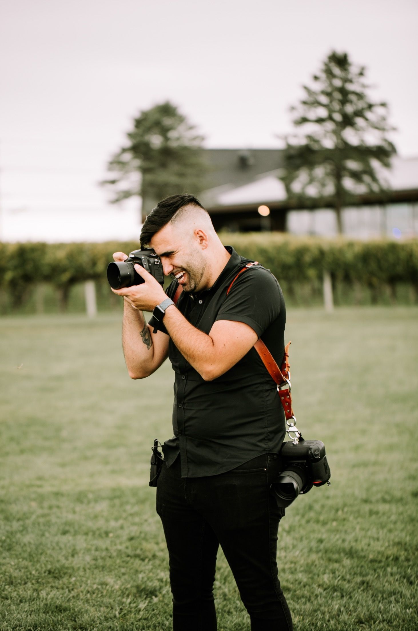 Wedding photographer in black attire captures shots in vineyard setting.