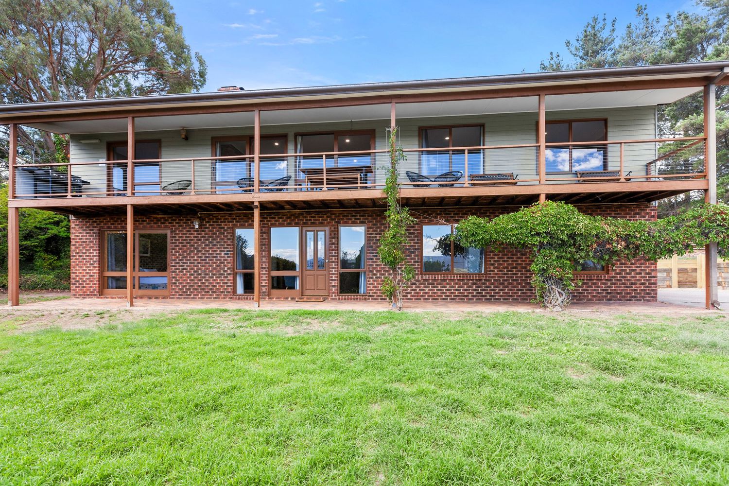 Modern two-story brick house with glass balcony overlooking a green lawn surrounded by trees.